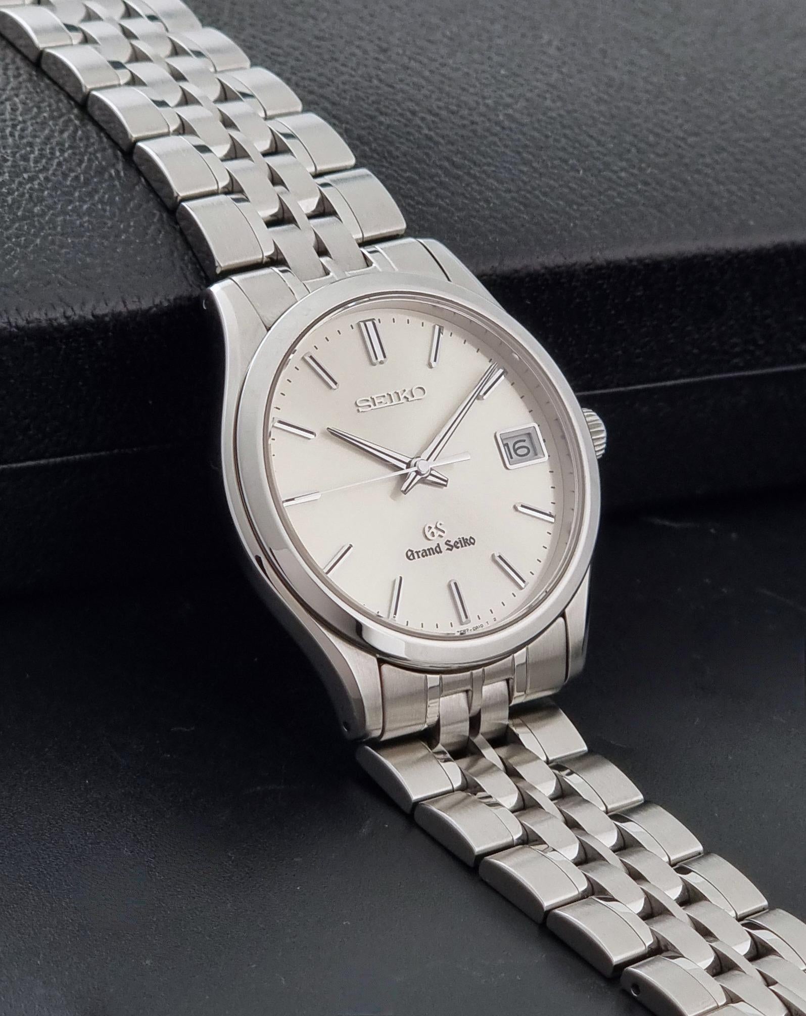 A Grand Seiko SBGV001 quartz watch and link bracelet and a cream-colored dial, resting on a textured black surface.