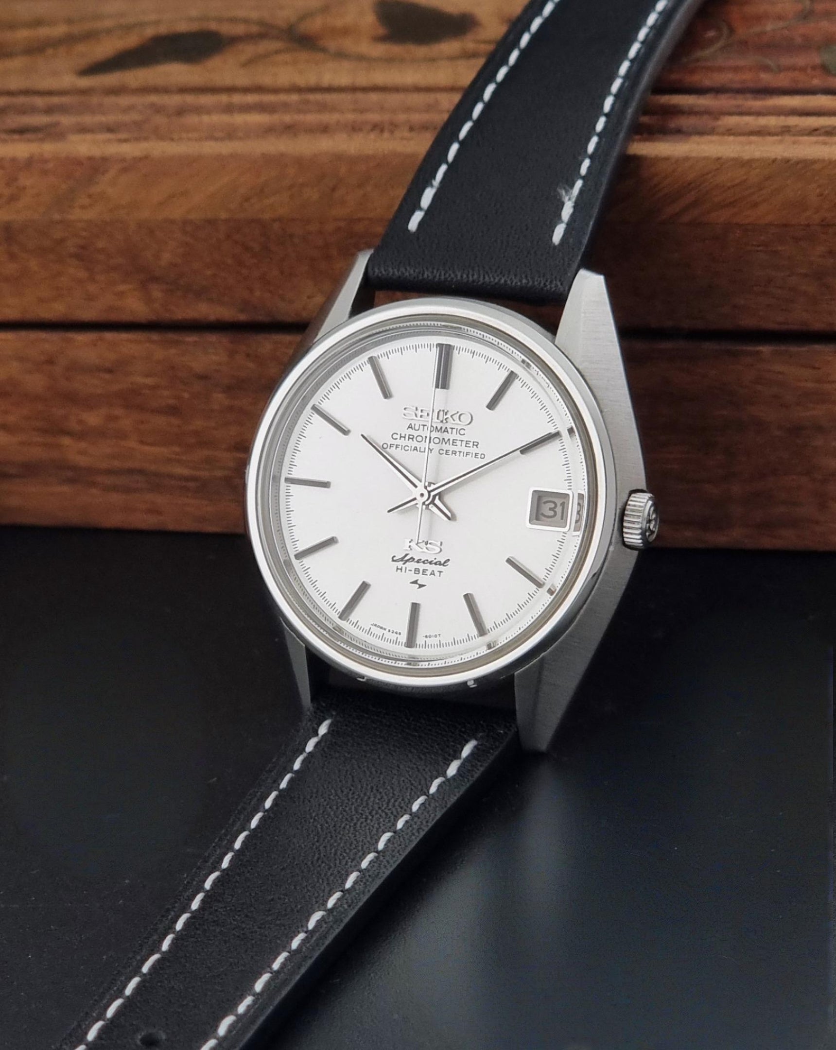 A Vintage King Seiko Special 5245-6010 Watch, white dial, and black leather strap with white stitching, photographed resting on a dark surface with a wooden background.