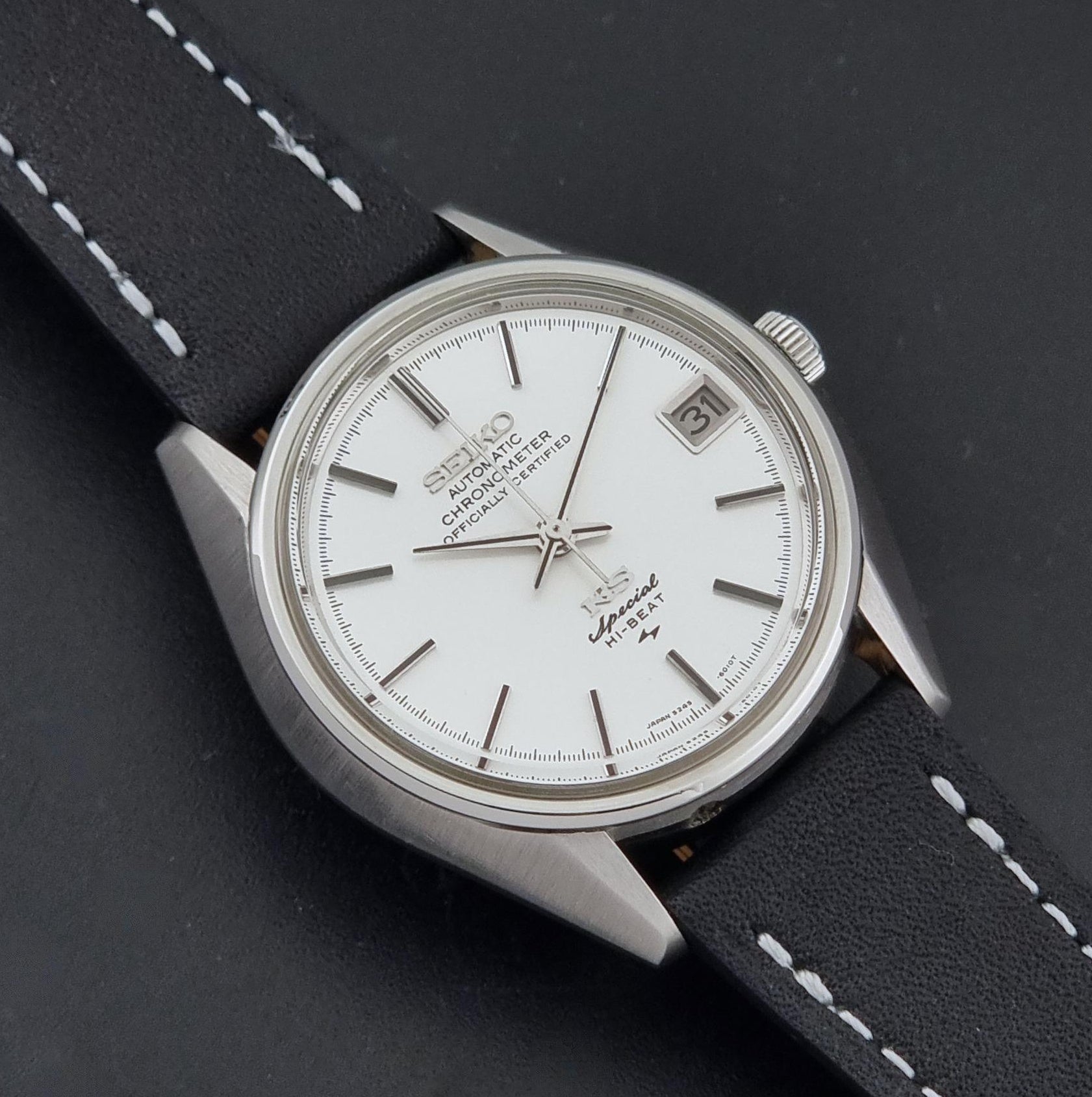 A Vintage King Seiko Special 5245-6010 Watch, white dial, and black leather strap with white stitching, photographed as a close up, shown on a dark textured background.
