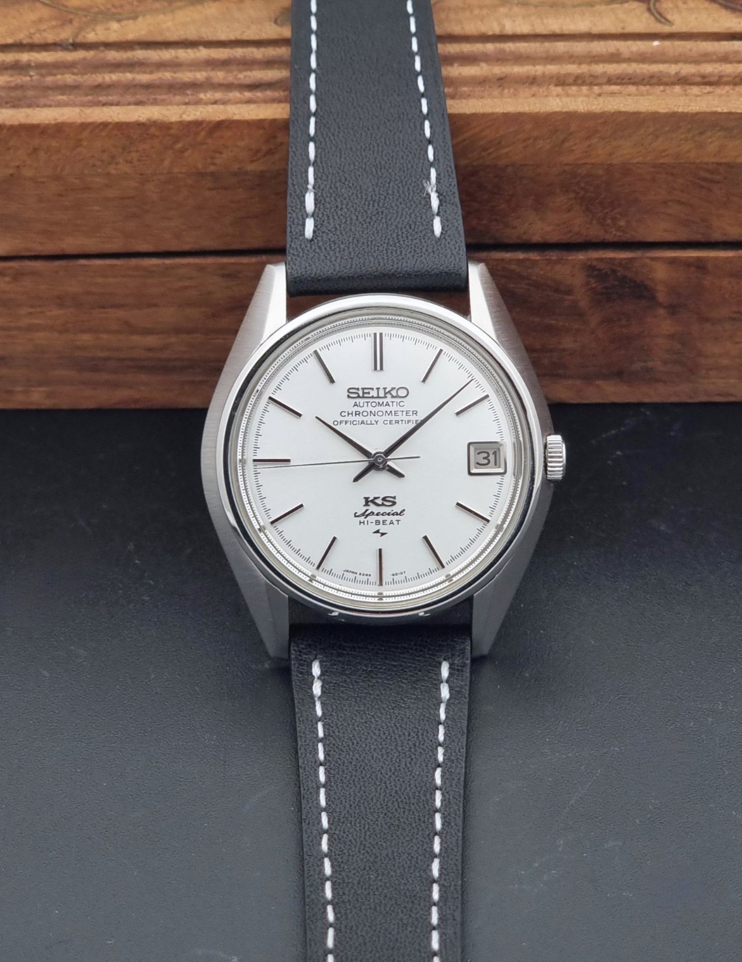 A Vintage King Seiko Special 5245-6010 Watch, white dial, and black leather strap with white stitching, photographed resting on a dark surface with a wooden background.