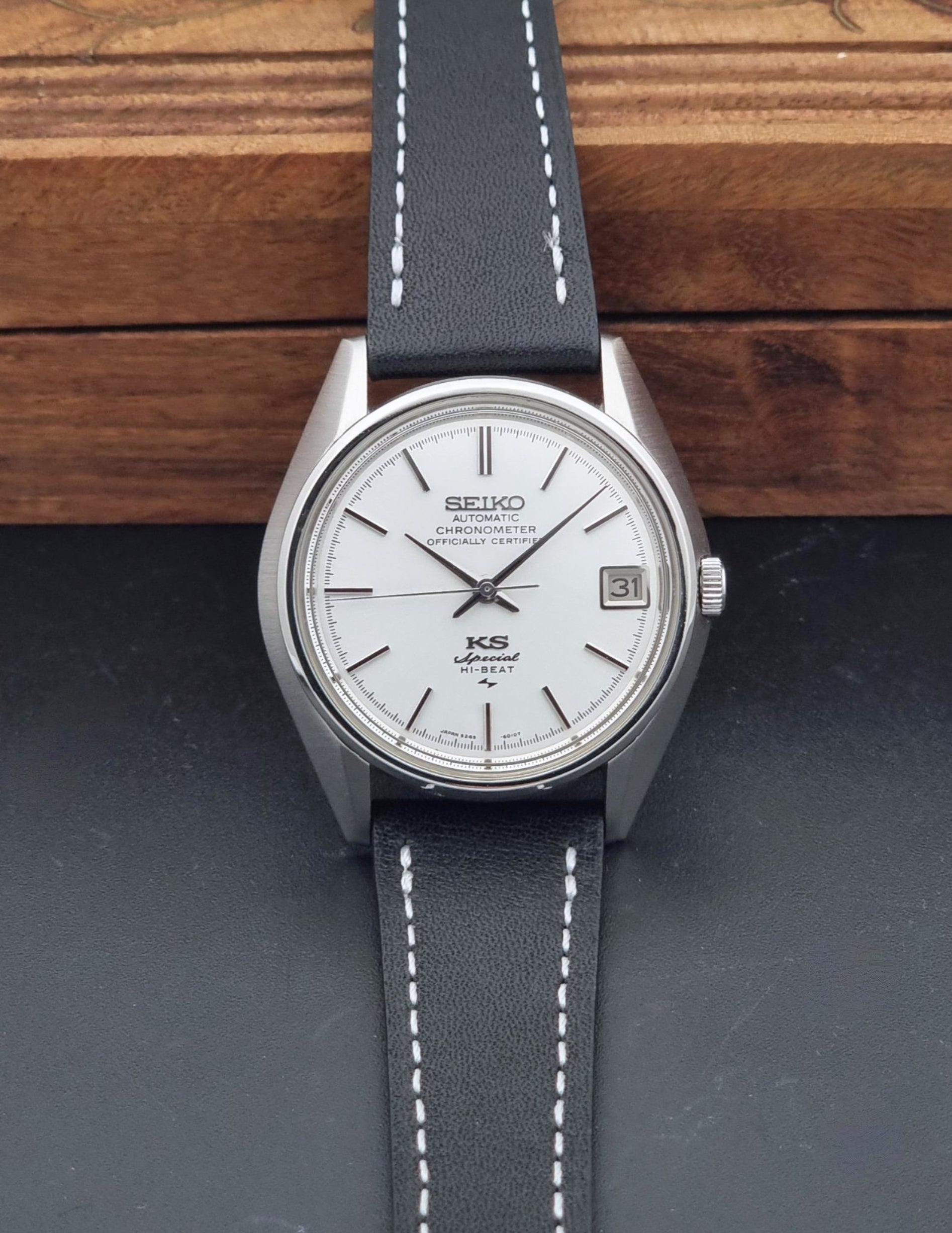A Vintage King Seiko Special 5245-6010 Watch, white dial, and black leather strap with white stitching, photographed resting on a dark surface with a wooden background.