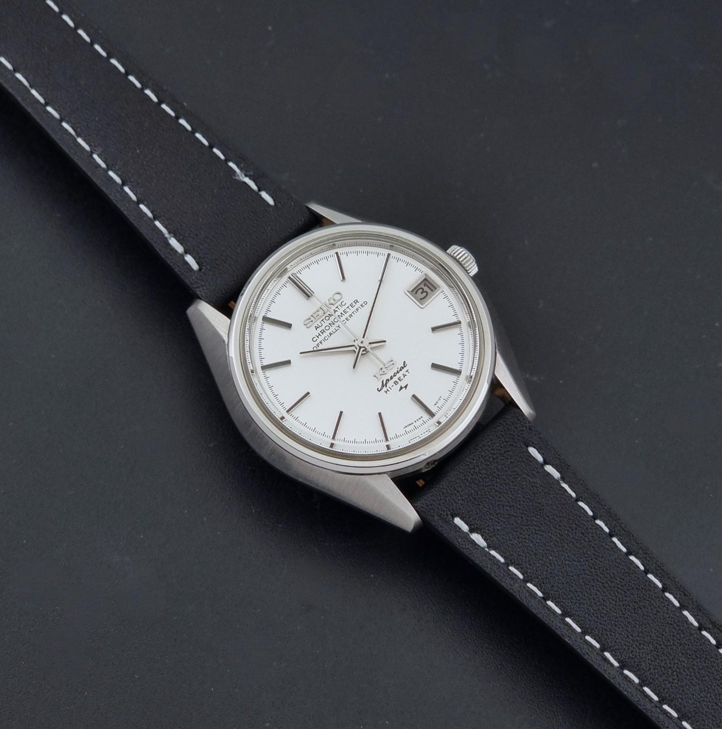 A Vintage King Seiko Special 5245-6010 Watch, white dial, and black leather strap with white stitching, photographed diagonally on a dark textured background.