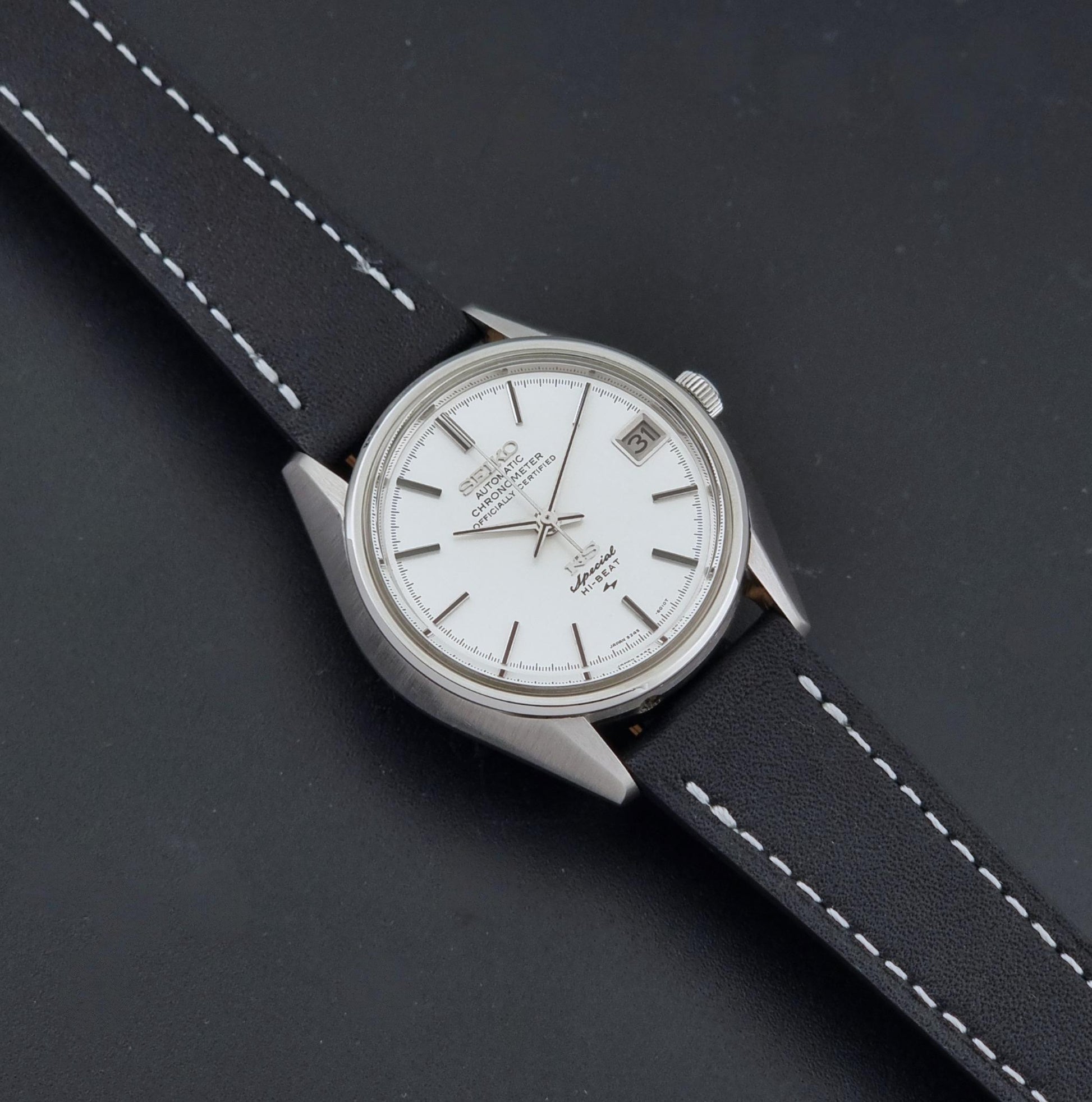 A Vintage King Seiko Special 5245-6010 Watch, white dial, and black leather strap with white stitching, photographed diagonally on a dark textured background.