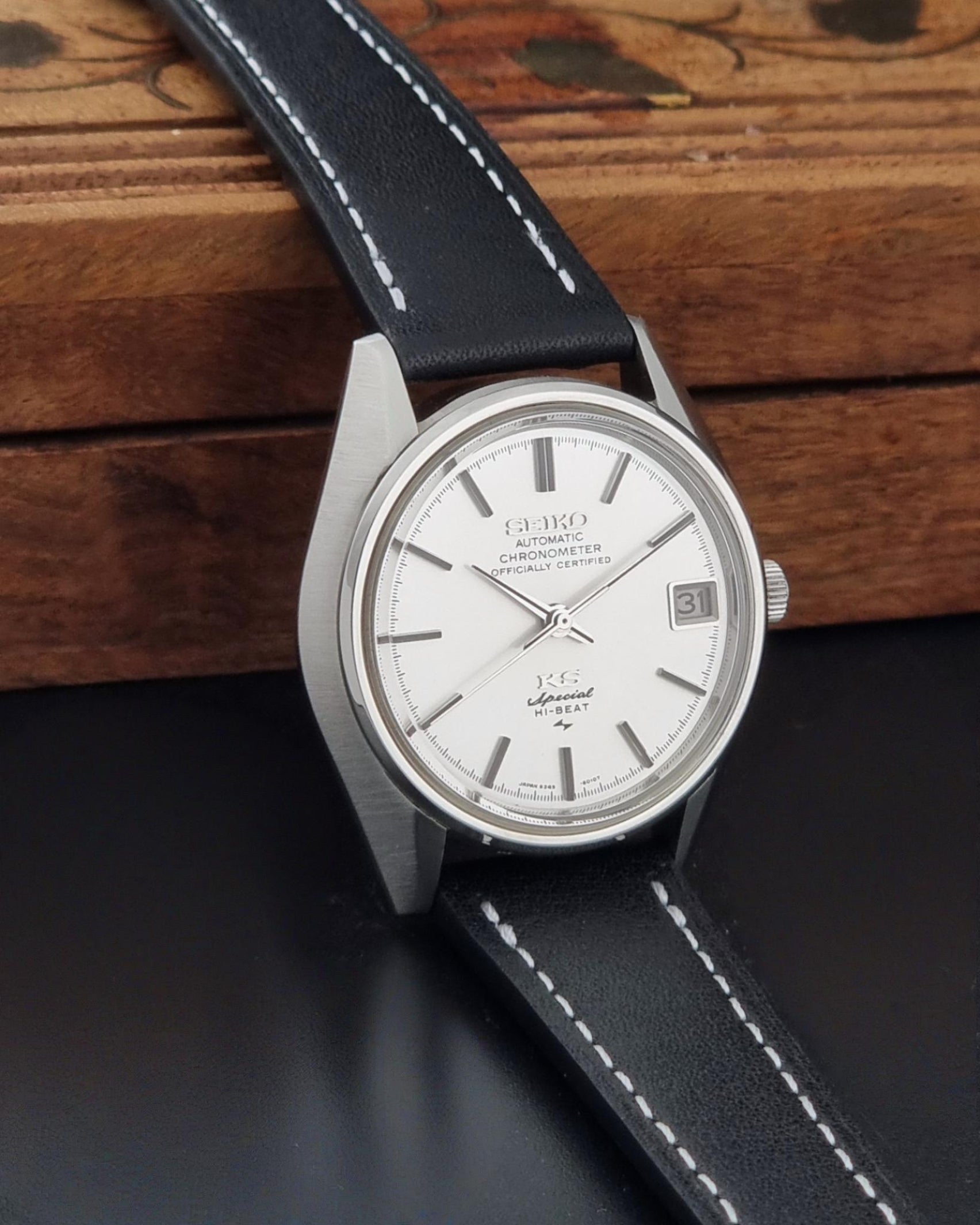 A Vintage King Seiko Special 5245-6010 Watch, white dial, and black leather strap with white stitching, photographed resting on a dark surface in front of a wooden box.