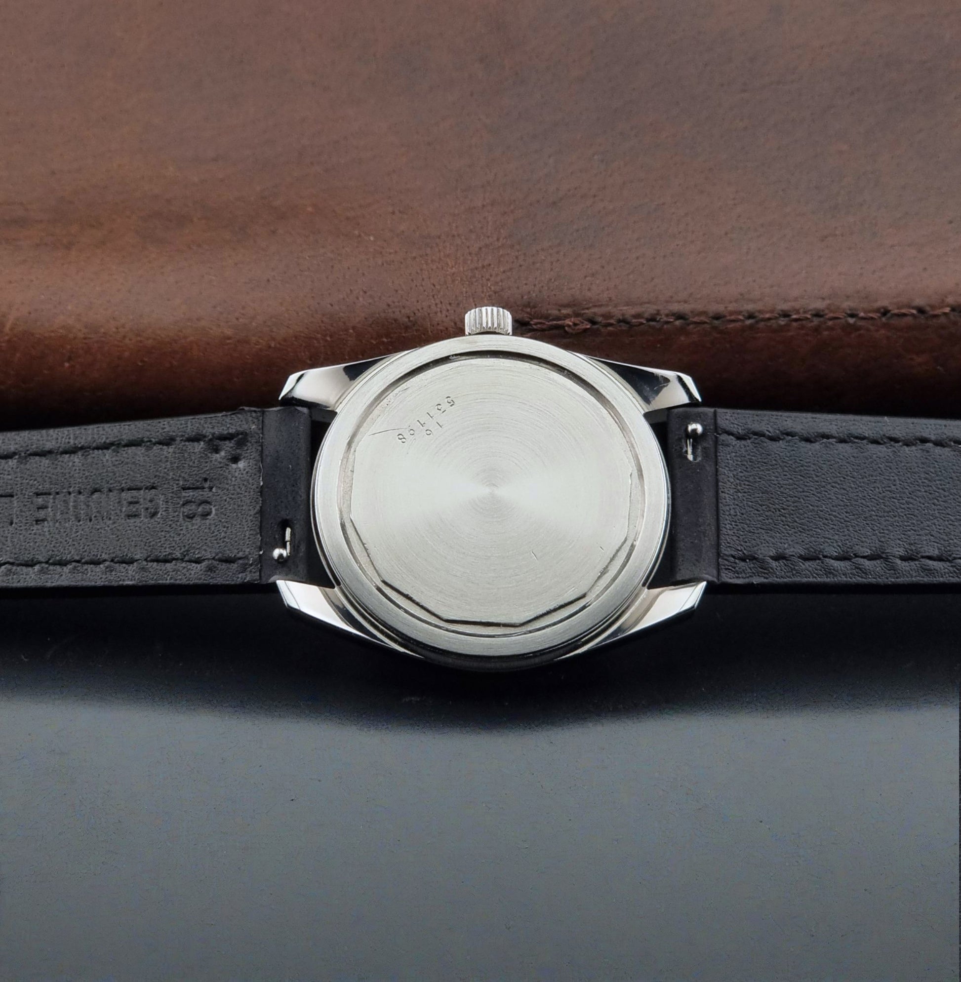 Close-up of the back of A Vintage Longines Admiral HF 2302 Wristwatch and black leather strap, photographed, resting on a dark surface with a brown leather background.