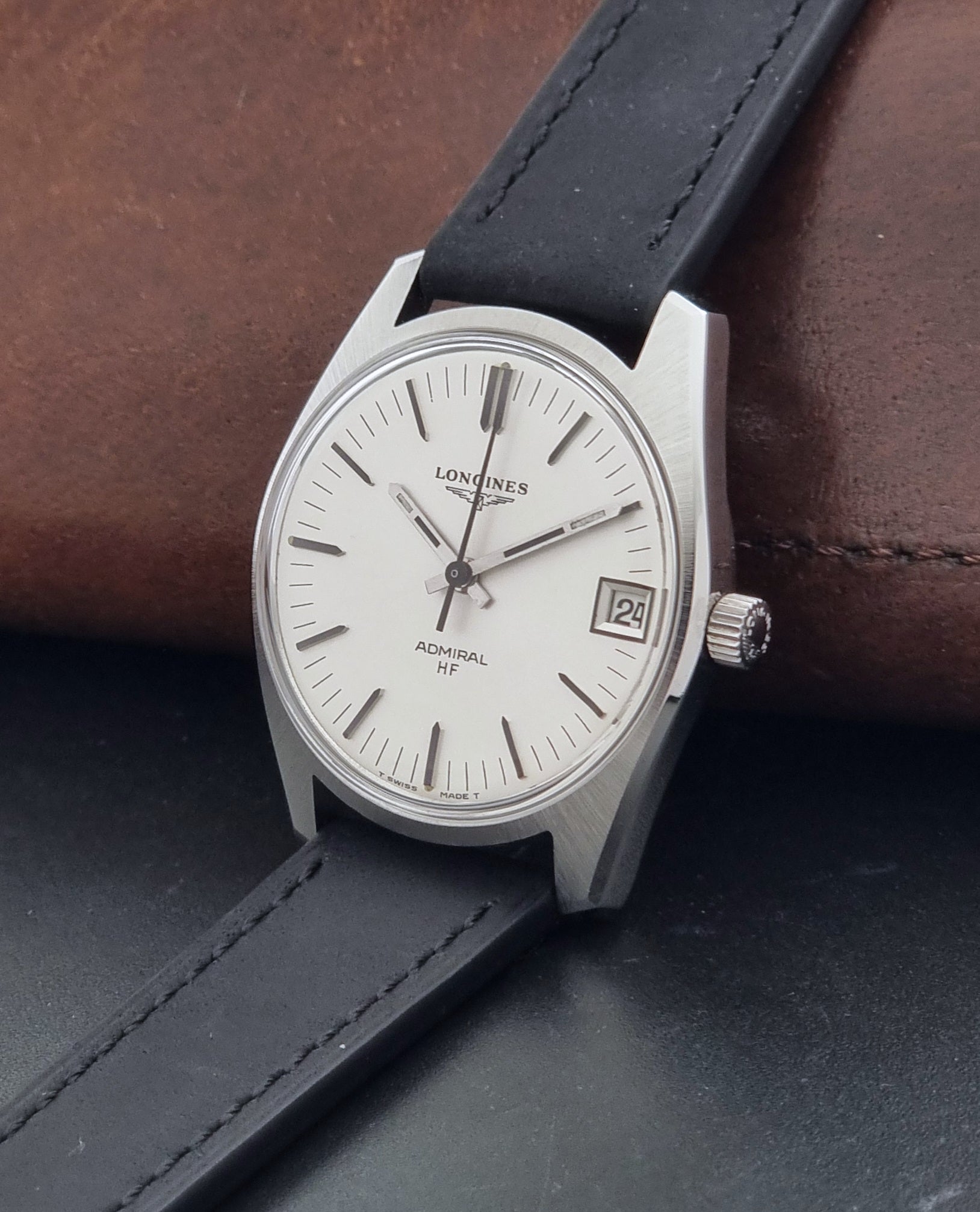 A Vintage Longines Admiral HF 2302 Wristwatch and black leather strap, photographed resting on a dark surface with a brown leather cylindrical prop behind it.
