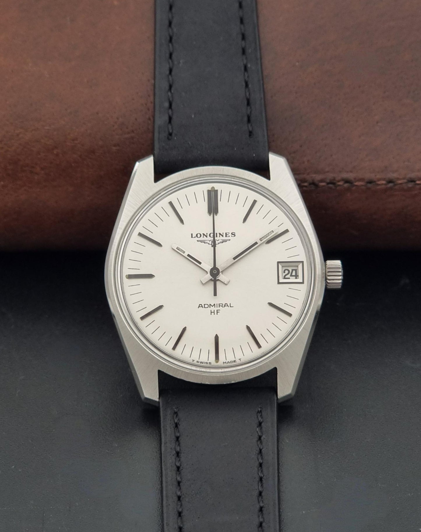 A Vintage Longines Admiral HF 2302 Wristwatch and black leather strap, displayed on a dark surface with a brown leather prop behind it.
