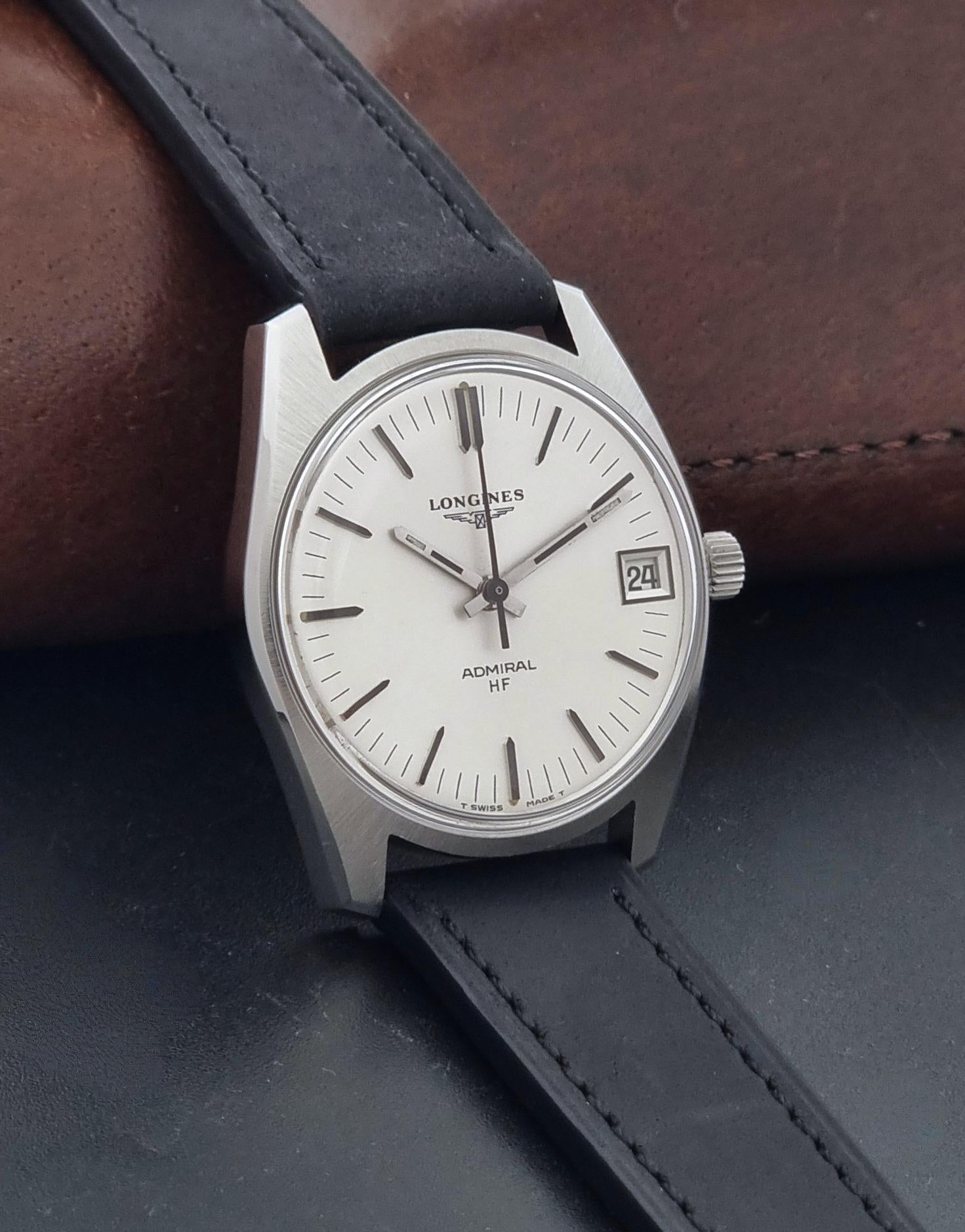 A Vintage Longines Admiral HF 2302 Wristwatch and black leather strap, photographed, resting on a dark surface with a brown leather prop in the background.