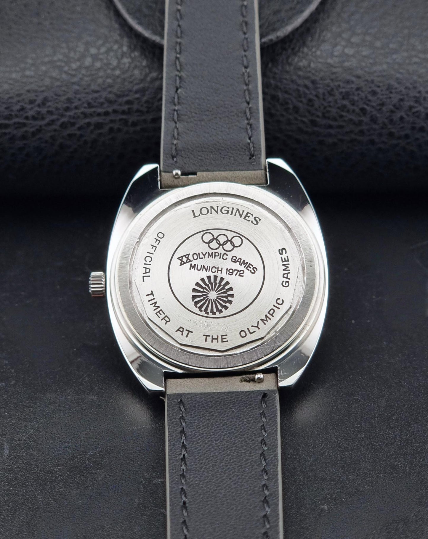 Close-up of the back of a Longines wristwatch with a silver-toned case and dark gray leather strap, showing the engraved 1976 Munich Olympic Games logo and text on a dark textured background.