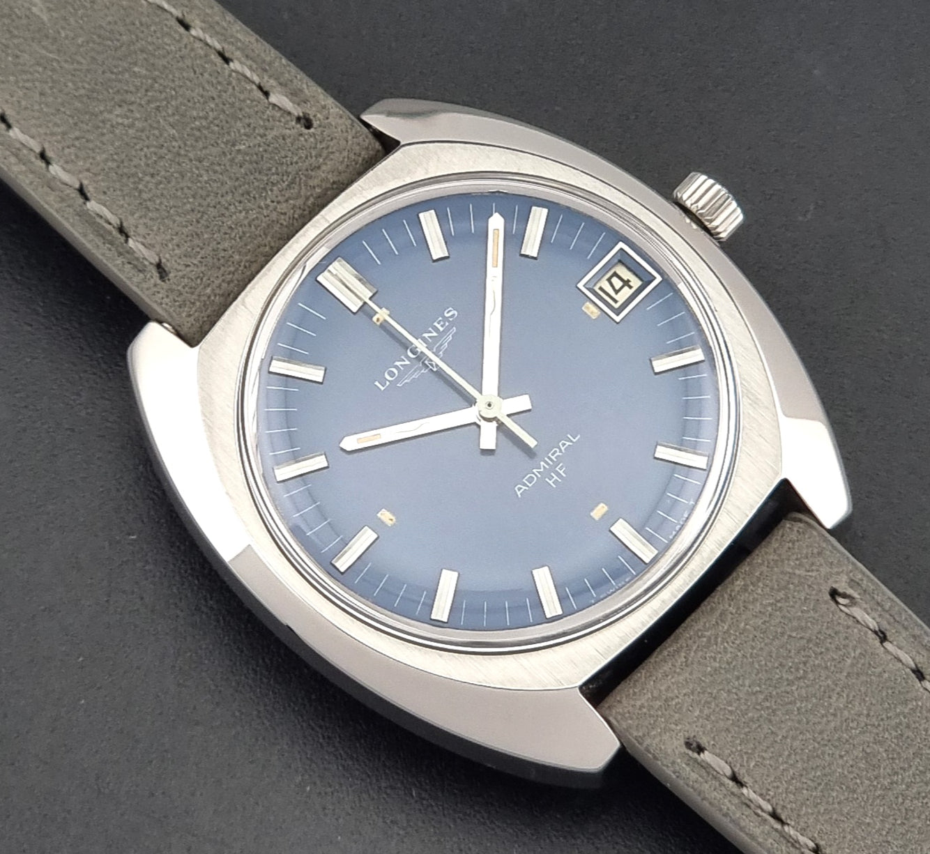 A close-up of a Longines Admiral HF wristwatch featuring a  blue dial, silver-toned stainless steel case, and a grey leather strap on a dark textured background.