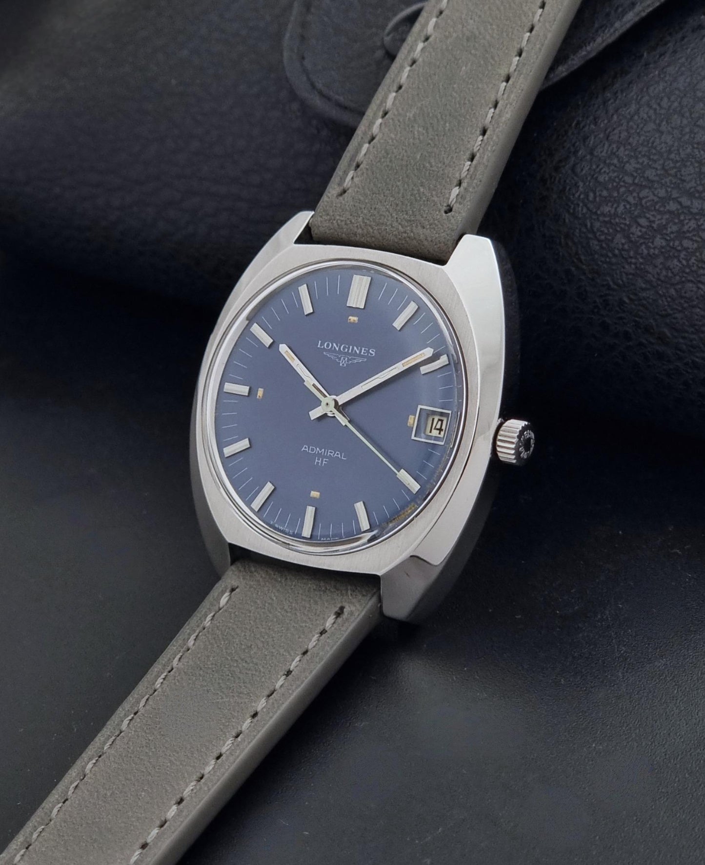 A Vintage Longines Admiral HF 2302 Wristwatch with a silver-toned case, blue dial, and gray leather strap, resting on a dark textured surface.