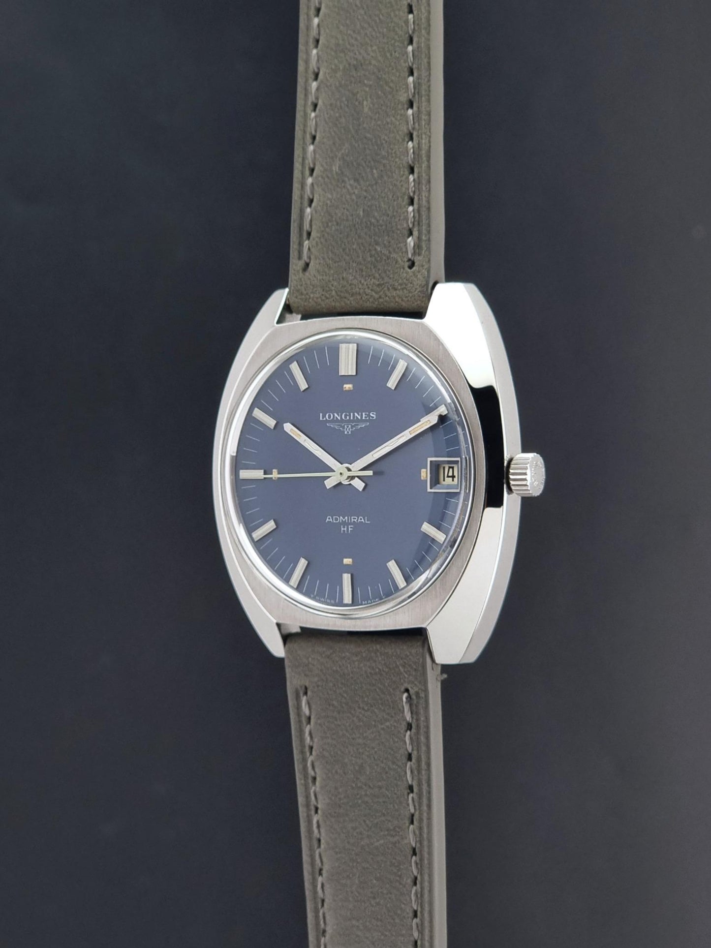 A vintage Longines watch with a silver-toned oval case, a blue dial, and a gray leather strap, shown against a dark gray background.