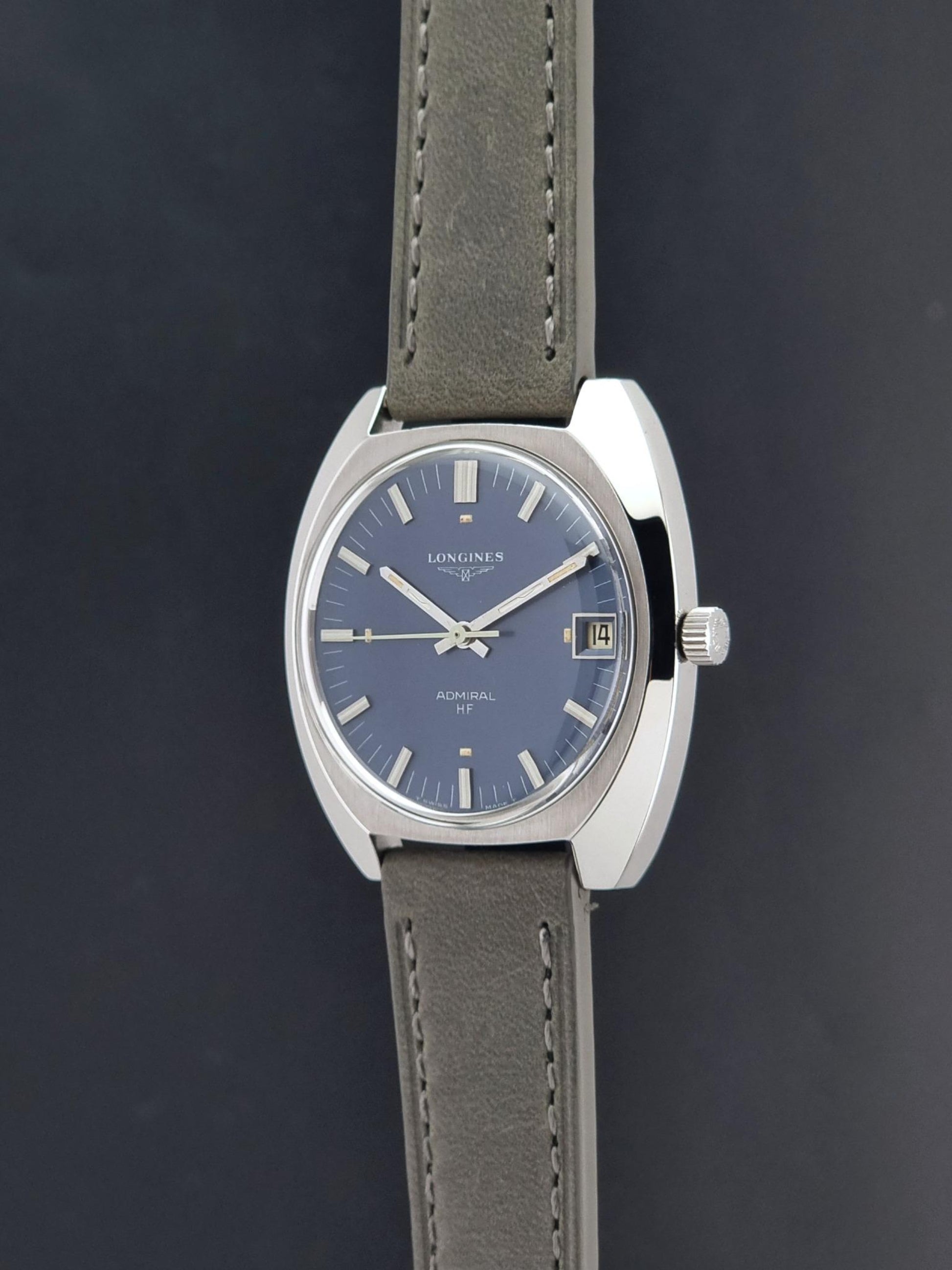 A vintage Longines watch with a silver-toned oval case, a blue dial, and a gray leather strap, shown against a dark gray background.