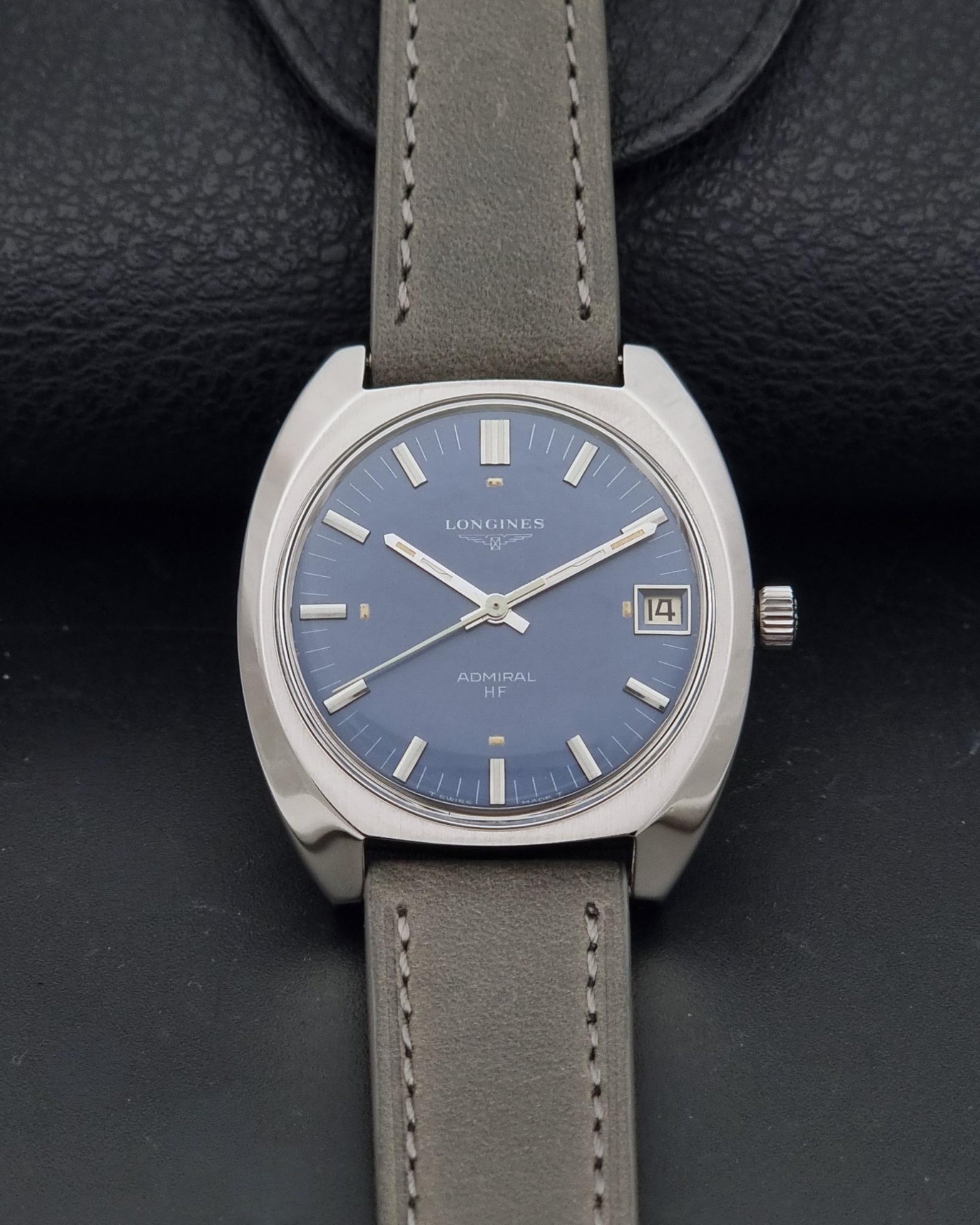A vintage Longines watch with a blue dial and a gray leather strap, displayed on a textured black background.