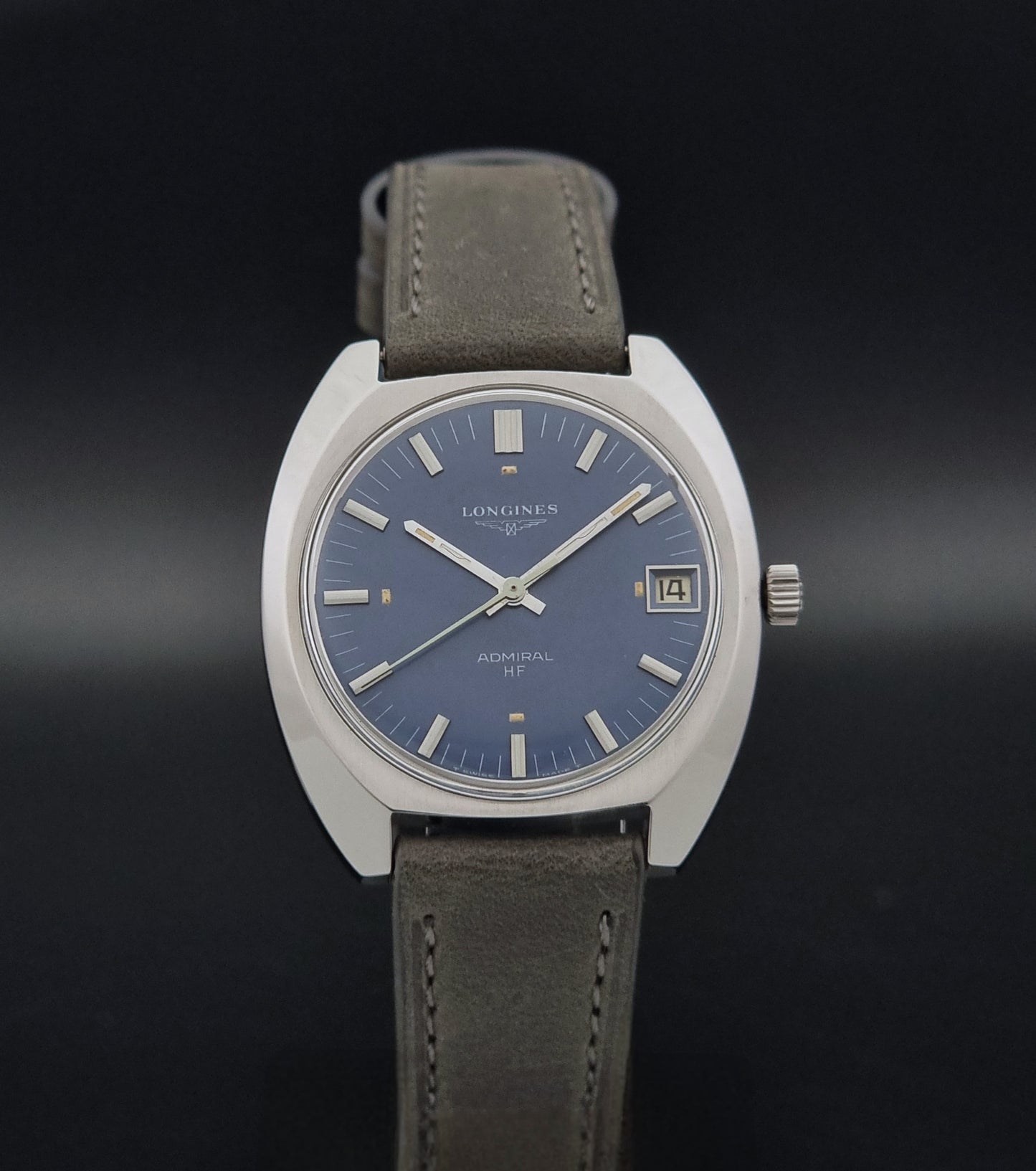 A Longines Admiral watch with a silver-toned round case, a blue dial, and and grey leather strap, shown against a dark gradient background.