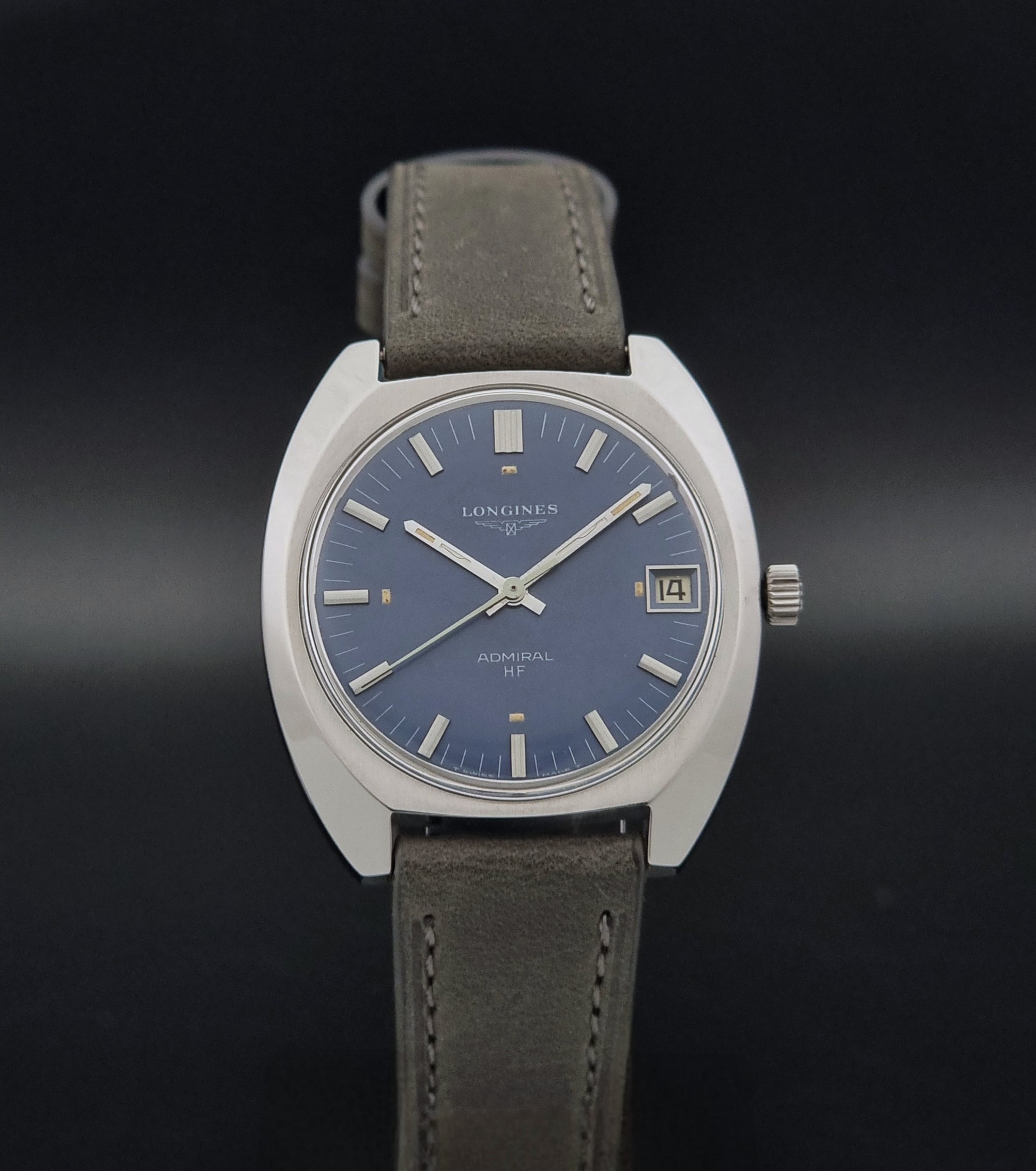 A Longines Admiral watch with a silver-toned round case, a blue dial, and and grey leather strap, shown against a dark gradient background.