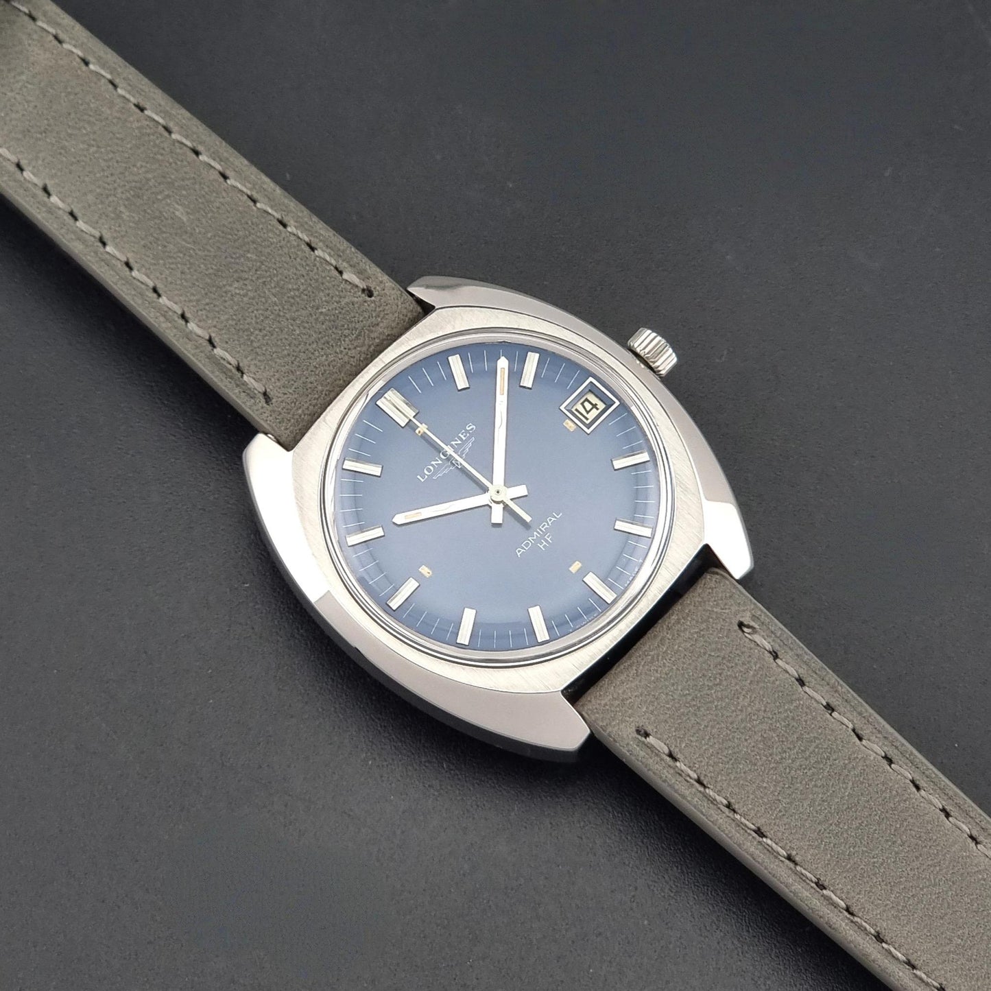 A Longines watch with a silver-toned stainless steel case, a dusty blue dial, and a taupe-brown leather strap, shown diagonally on a dark gray textured background.