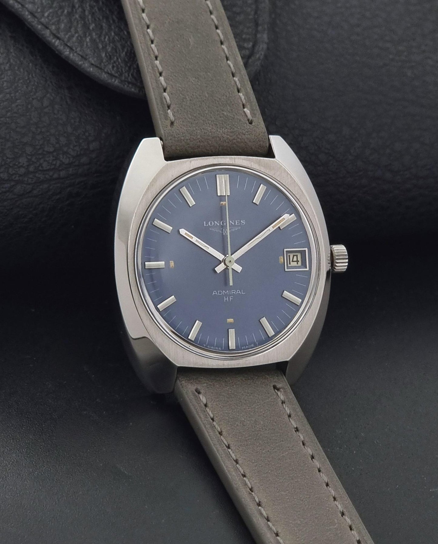 A Vintage Longines Admiral HF 2302 Wristwatch, a blue dial, and a grey leather strap, resting on a dark textured leather surface.