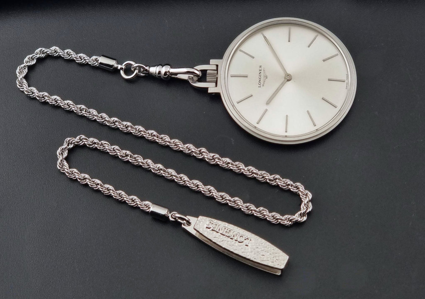 A silver-toned Longines 7713 pocket watch with a white dial and matching chain, displayed on a dark gray background. The chain includes a separate rectangular tag engraved with the brand name 'LONGINES'.