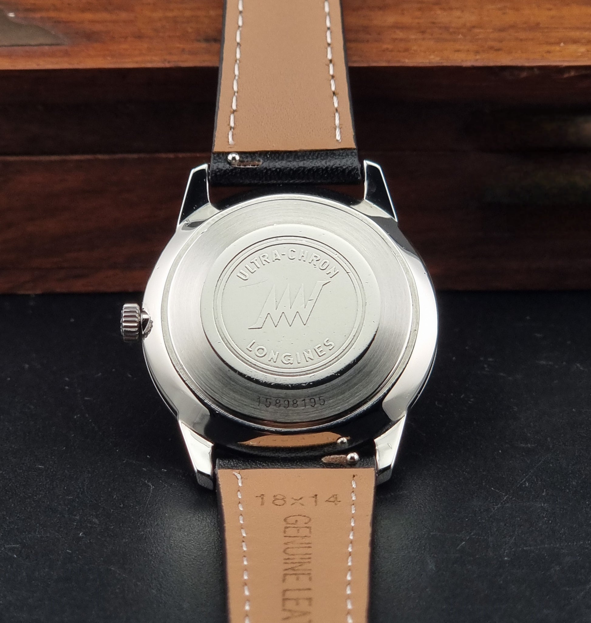 Close-up of the back of a Longines wristwatch with a silver metal case and a tan leather strap, resting on a dark surface with a wooden background.