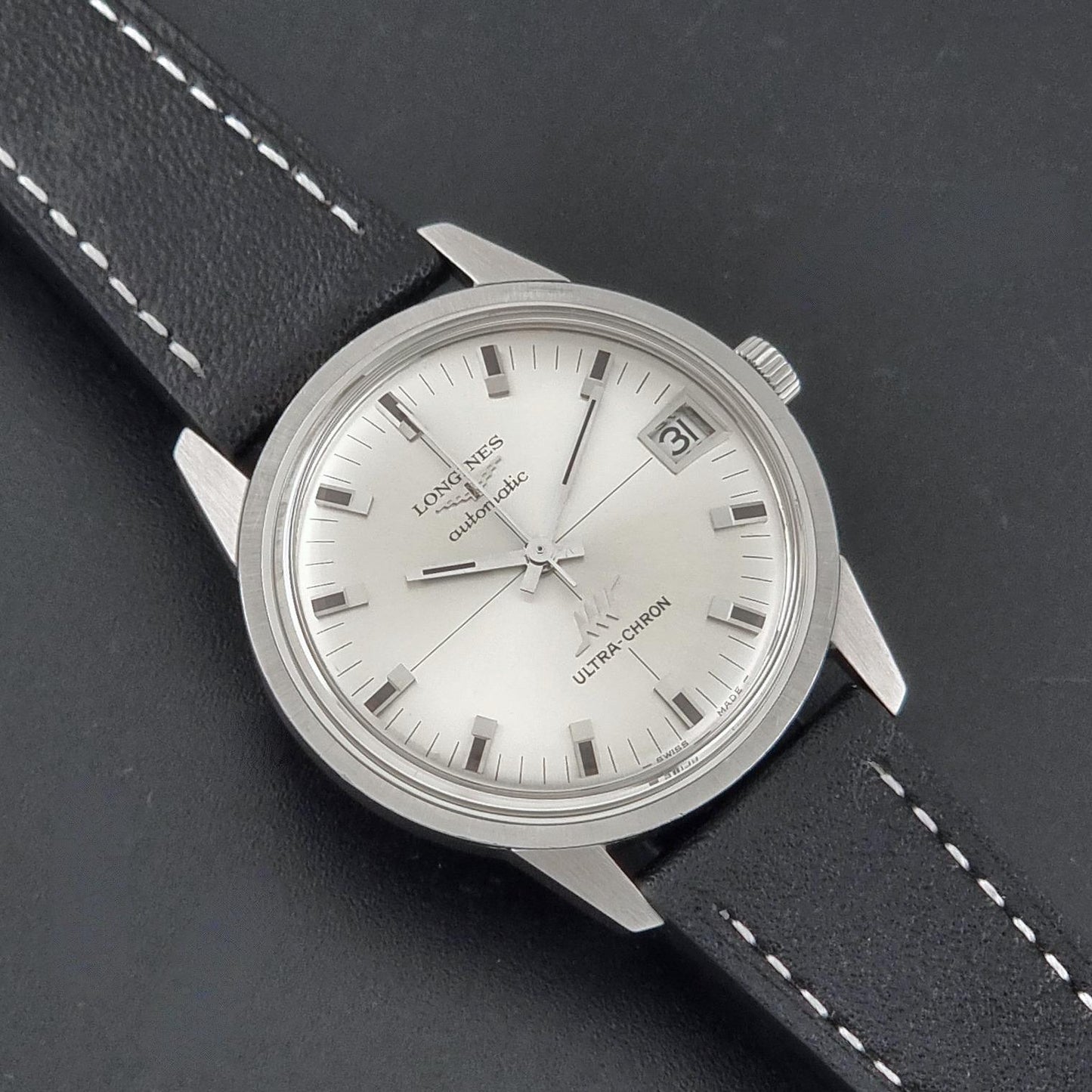 A close-up of a classic Vintage Longines ULTRA-CHRON 8309 watch with a silver-toned stainless steel case, a white dial with stick indices, and a black leather strap with white stitching, set against a dark textured background.