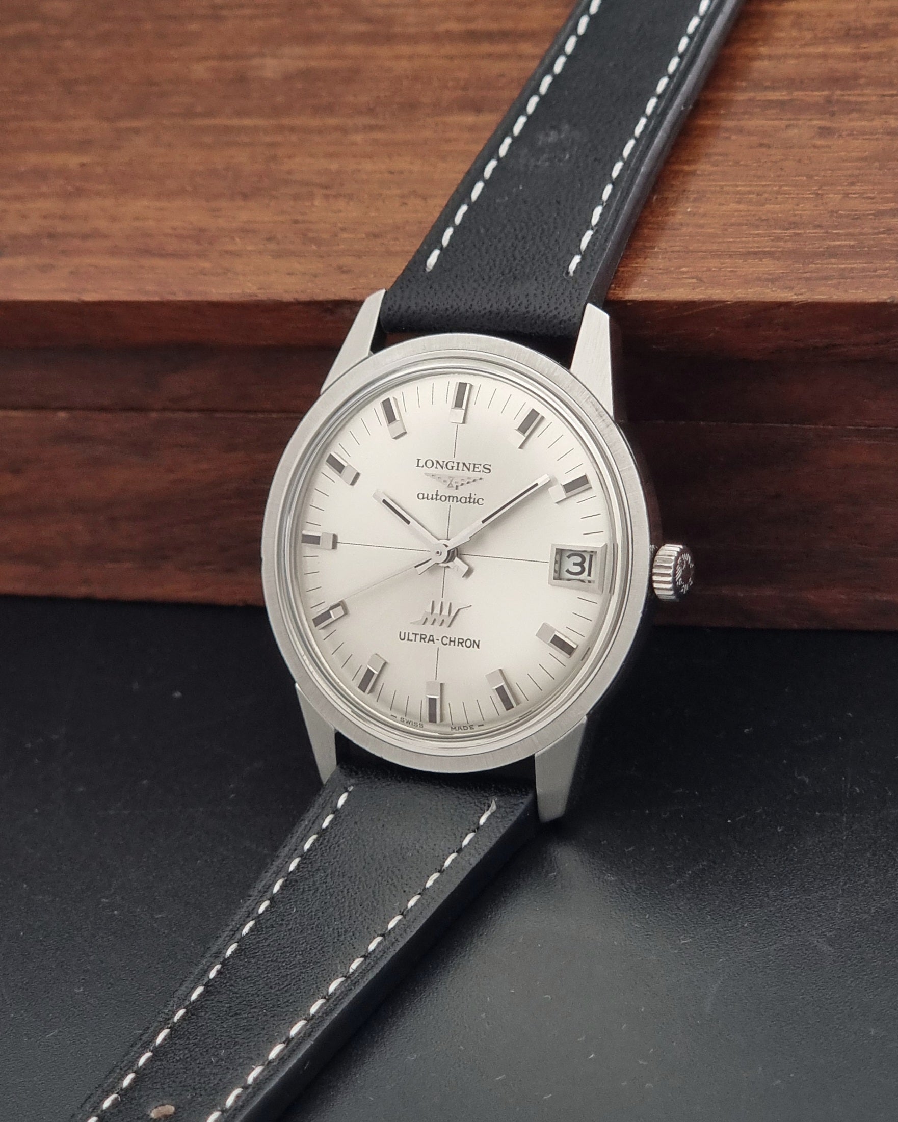 A Vintage Longines ULTRA-CHRON 8309 watch, white dial, and black leather strap with white stitching, resting on a dark surface with a wooden block in the background.