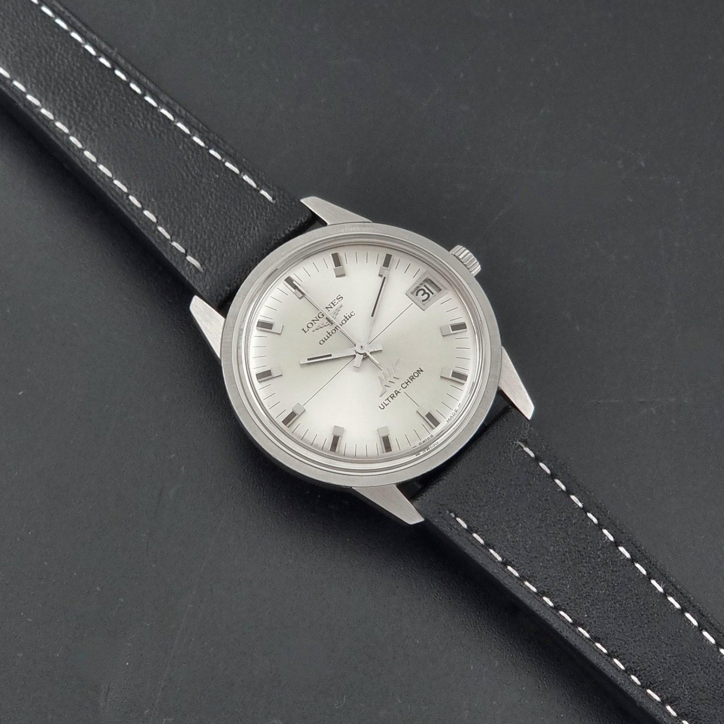 A Vintage Longines ULTRA-CHRON 8309 watch, white dial, and black leather strap with white stitching, photographed on a dark textured background.