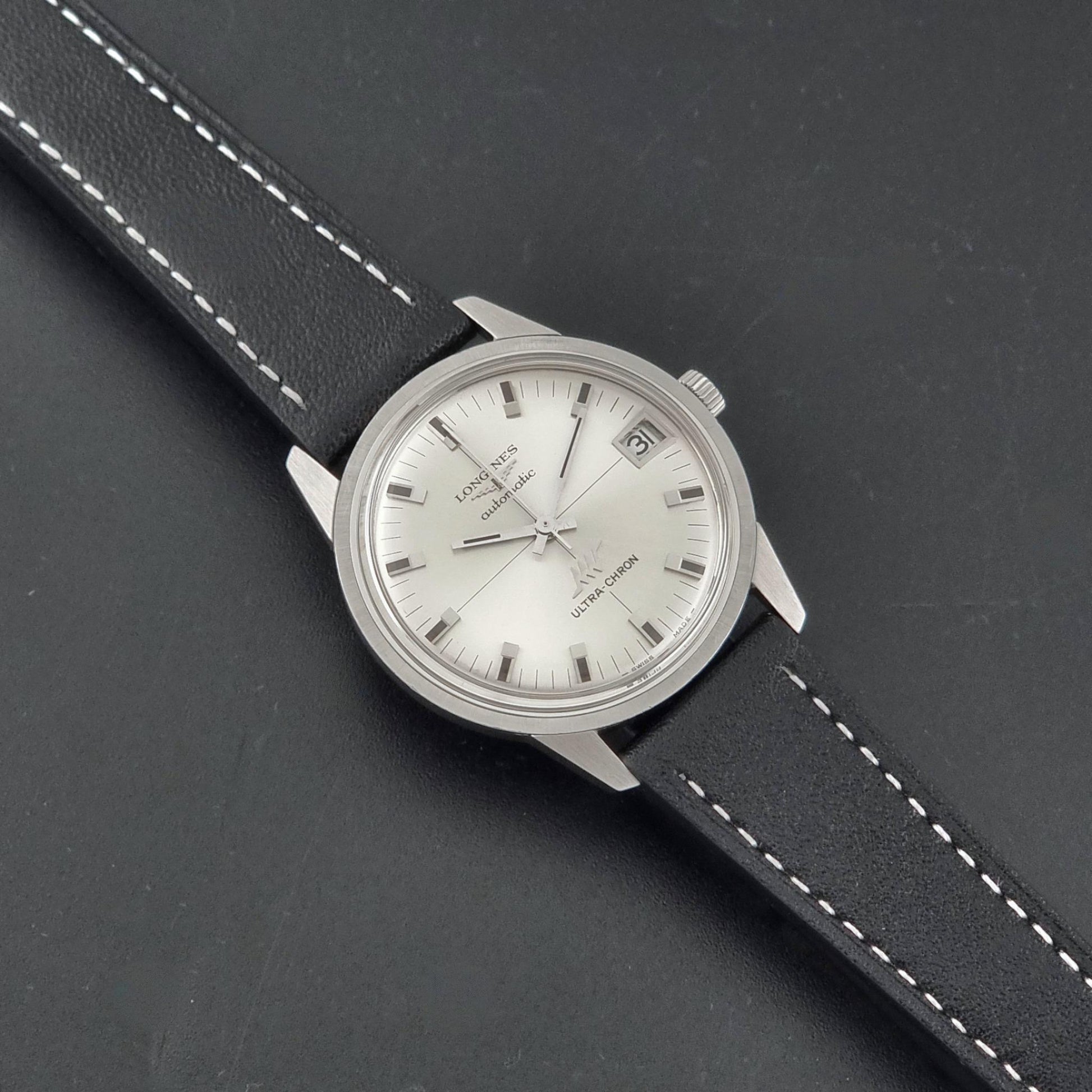 A Vintage Longines ULTRA-CHRON 8309 watch, white dial, and black leather strap with white stitching, photographed on a dark textured background.