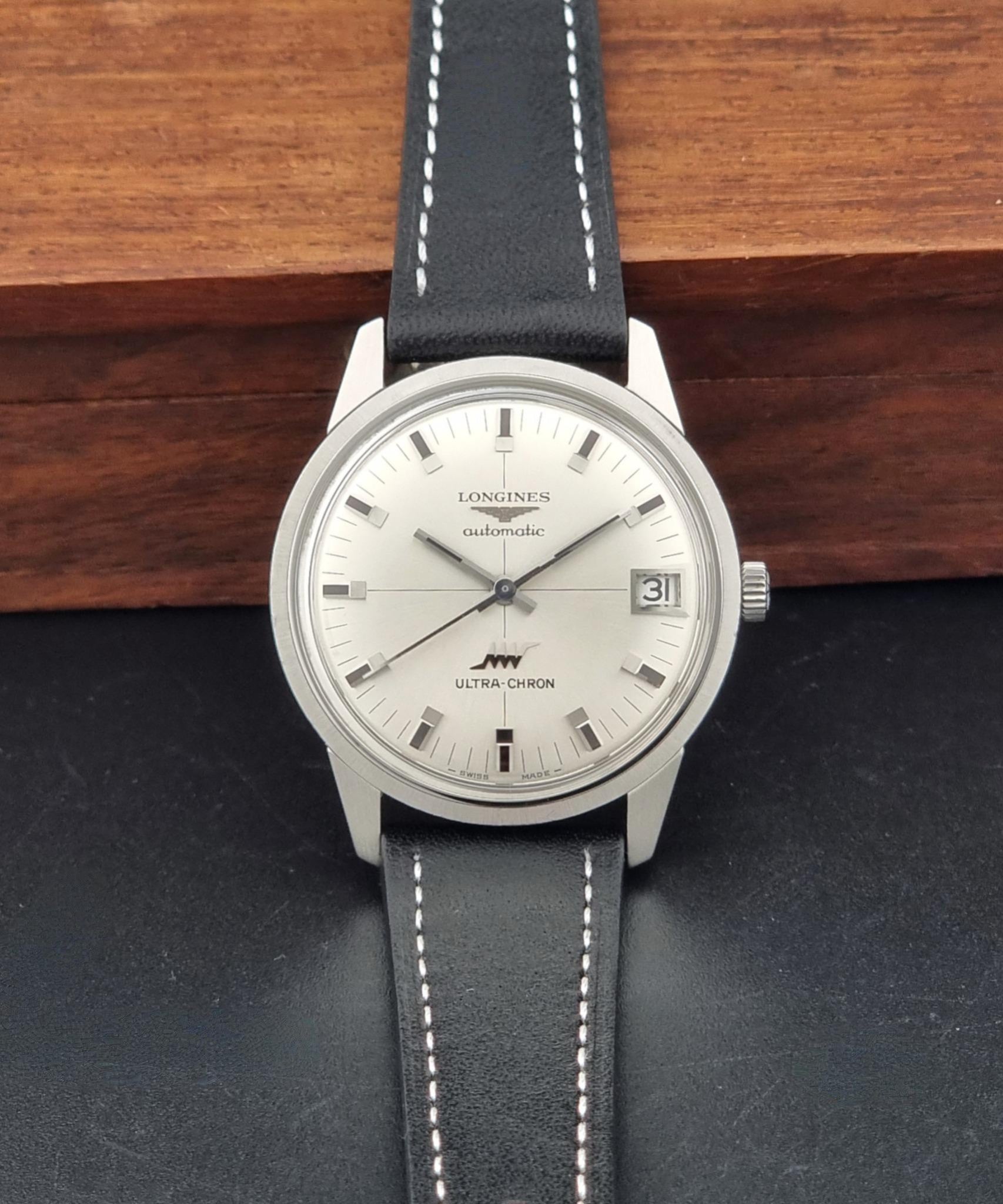A Longines Automatic watch with a silver-toned stainless steel case, white dial, and a black leather strap with white stitching, displayed against a dark surface and a wooden background.