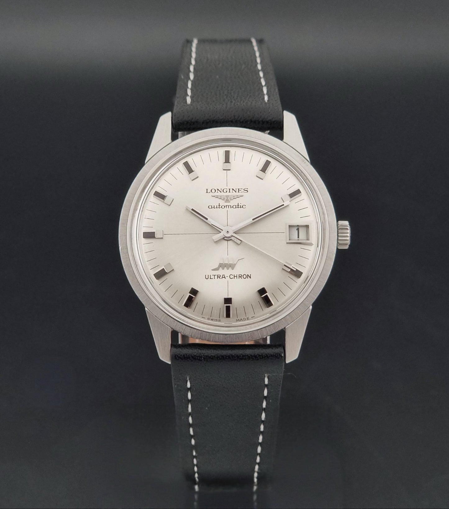 A Longines Ultra-Chron automatic watch with a silver-toned stainless steel case, white dial, and a black leather strap with white stitching, shown against a dark gray background.
