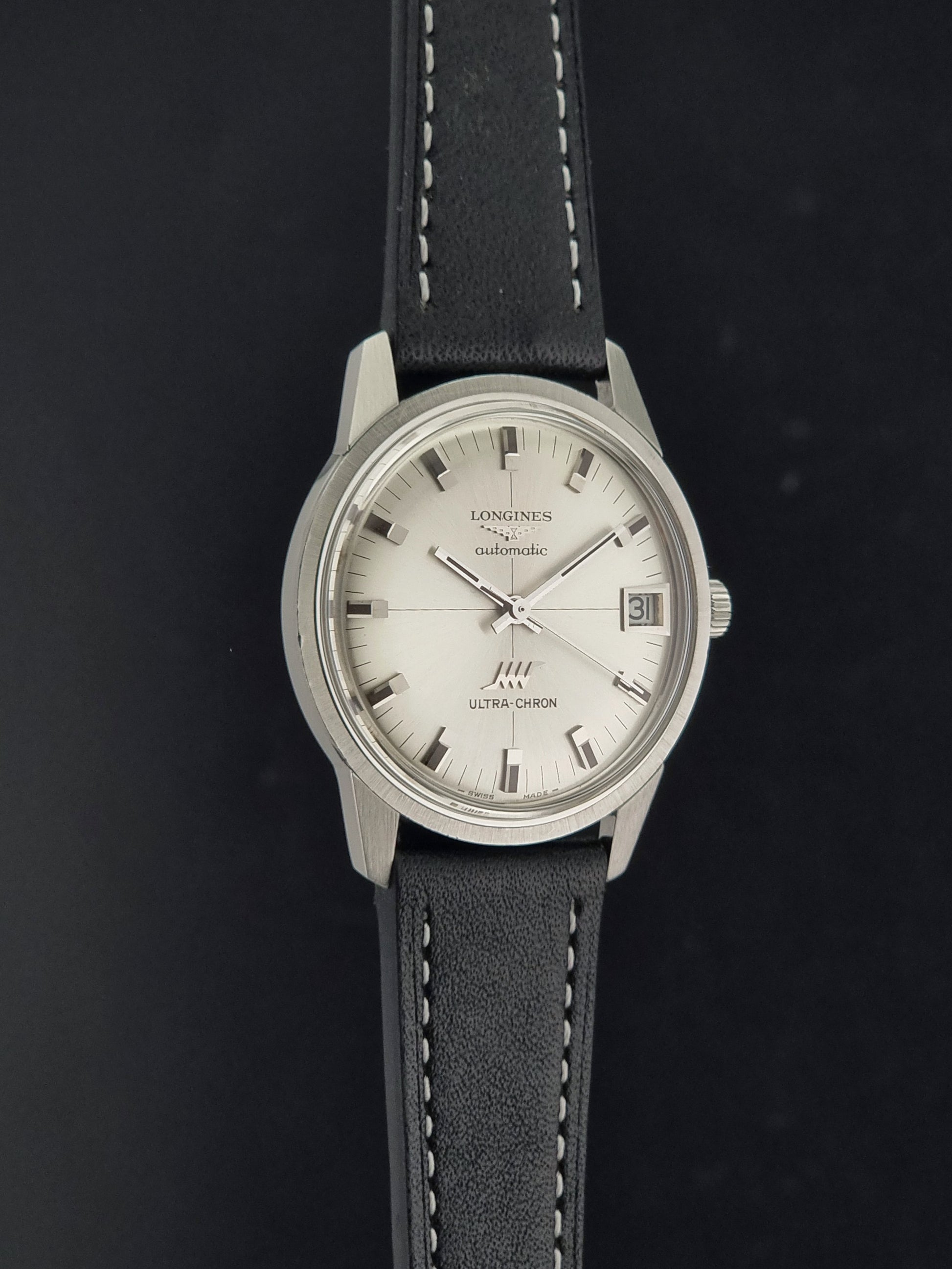 A Vintage Longines ULTRA-CHRON 8309 watch with a silver-toned round case, white dial, and black leather strap with white stitching, shown against a dark background.