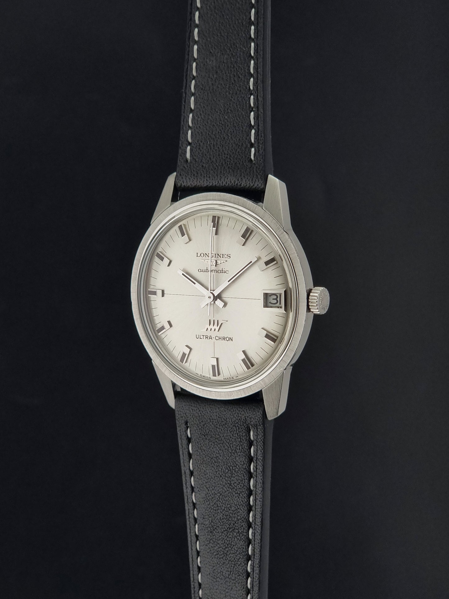 A Vintage Longines ULTRA-CHRON 8309 watch with a silver-toned metal case, a white dial, and a black leather strap with white stitching, shown against a dark background.