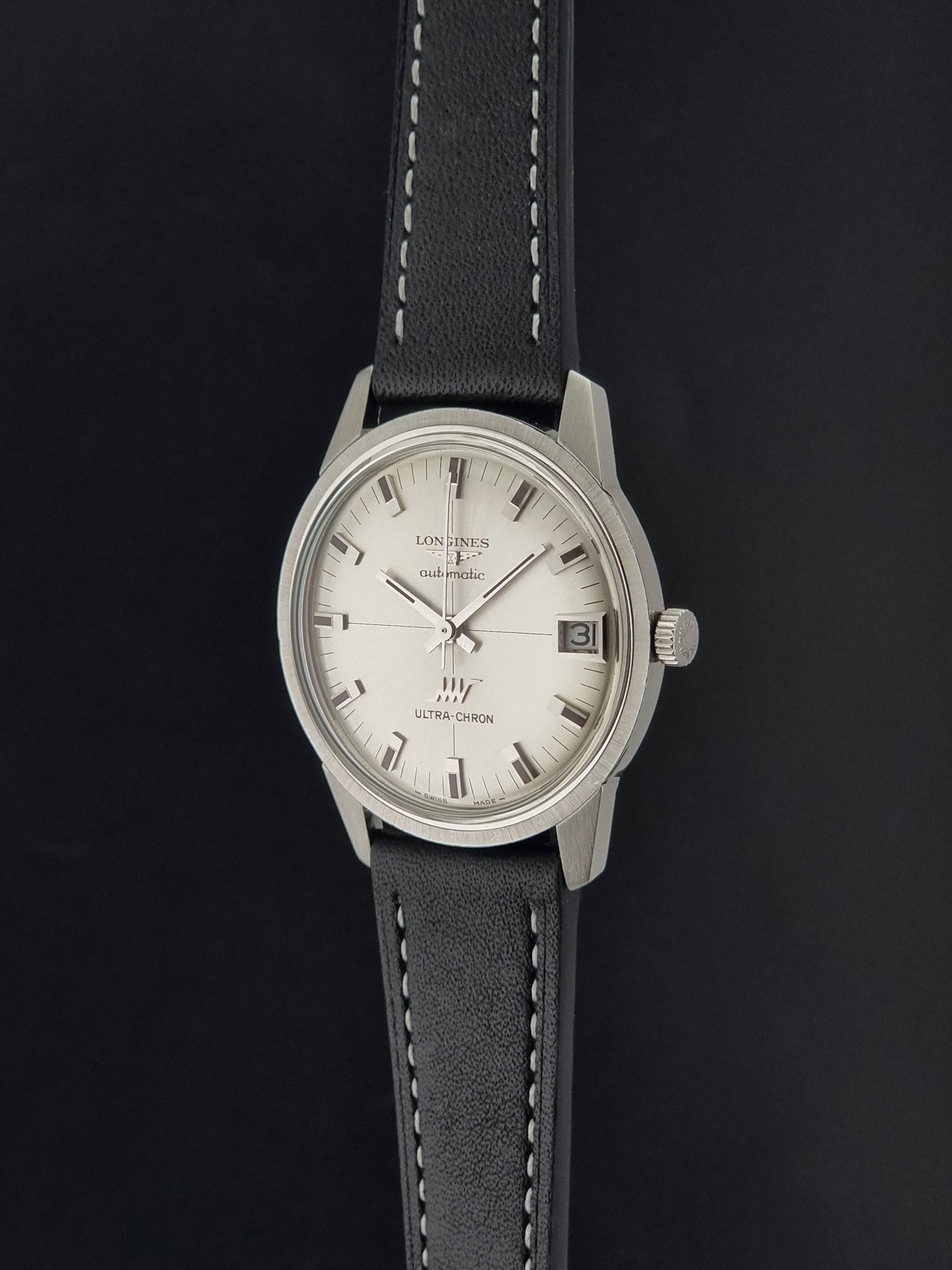 A Vintage Longines ULTRA-CHRON 8309 watch with a silver-toned metal case, a white dial, and a black leather strap with white stitching, shown against a dark background.