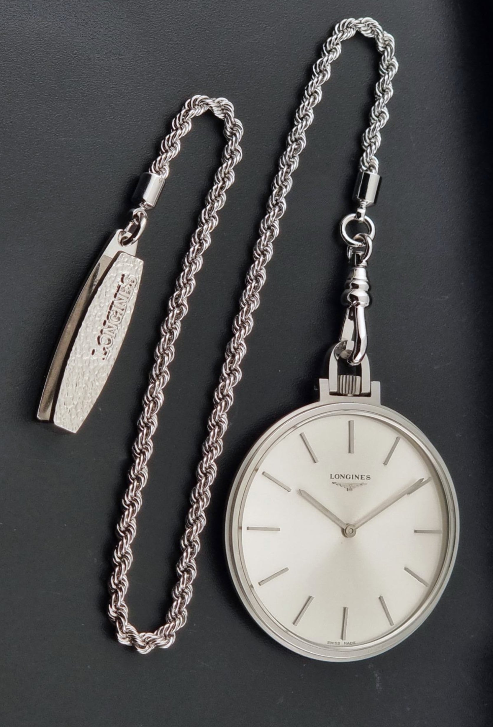 A silver-toned Longines pocket watch with a white dial, displayed alongside its matching braided chain and separate rectangular watch end piece on a dark gray background.