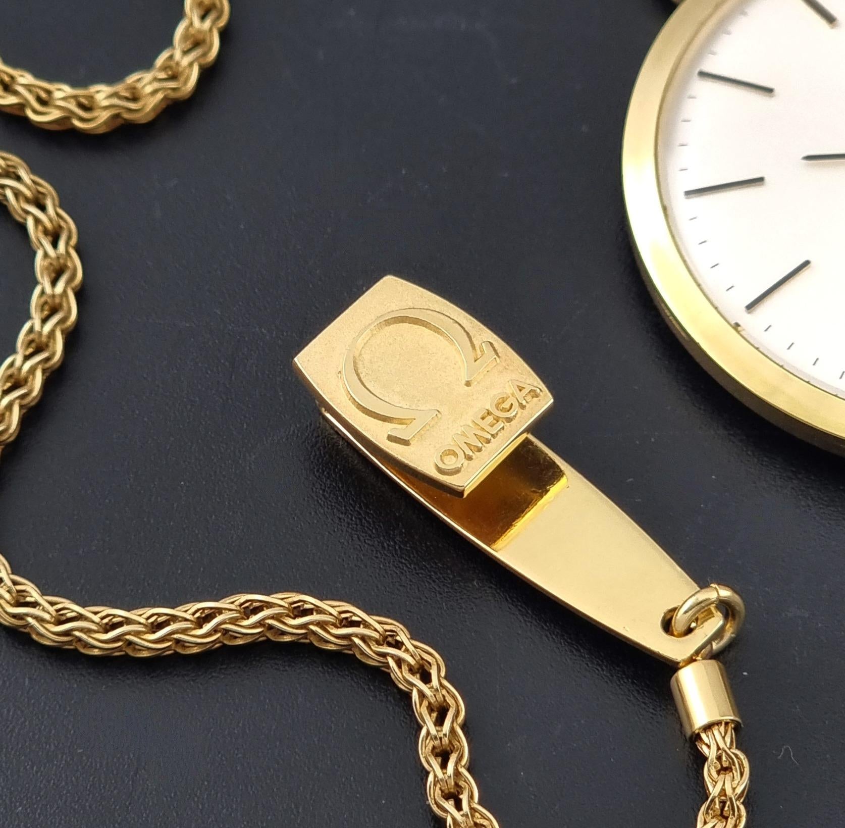 A gold-tone Omega watch chain with a polished rectangular tag engraved with the Omega logo, displayed on a dark textured surface next to a partial watch face.