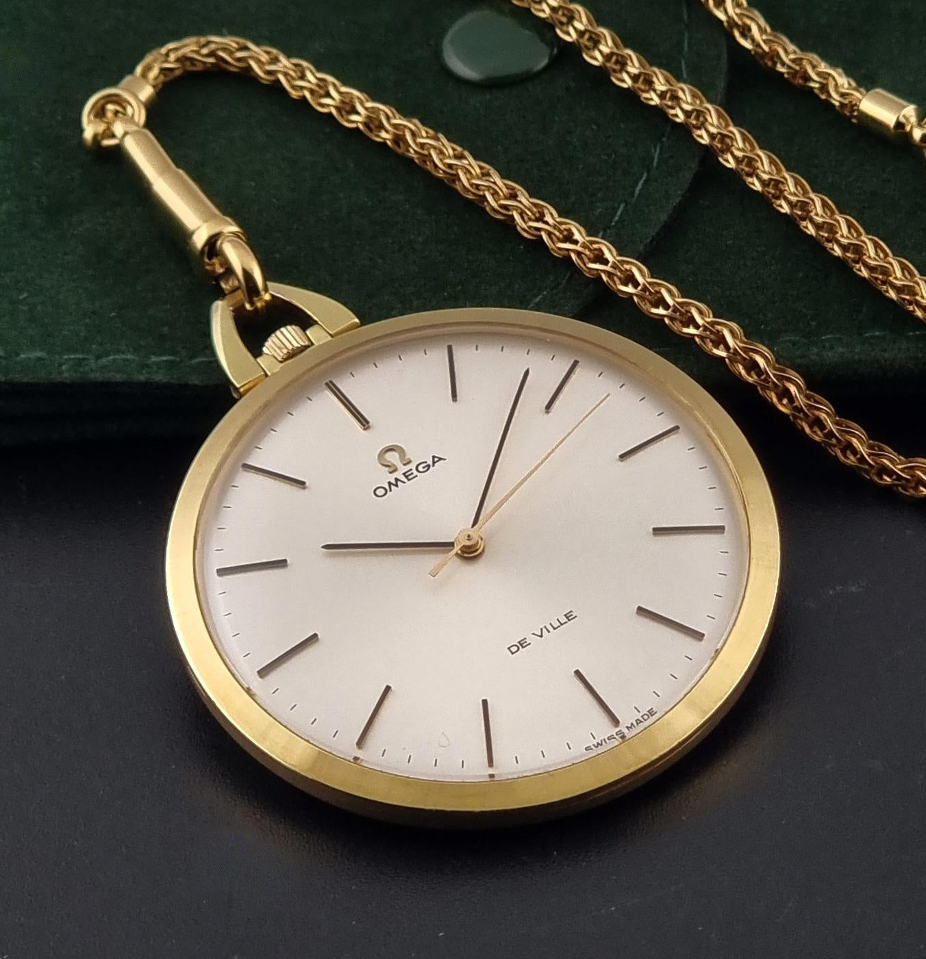 A gold Omega De Ville pocket watch with a white dial and matching gold chain, displayed on a dark surface with a green velvet pouch in the background.