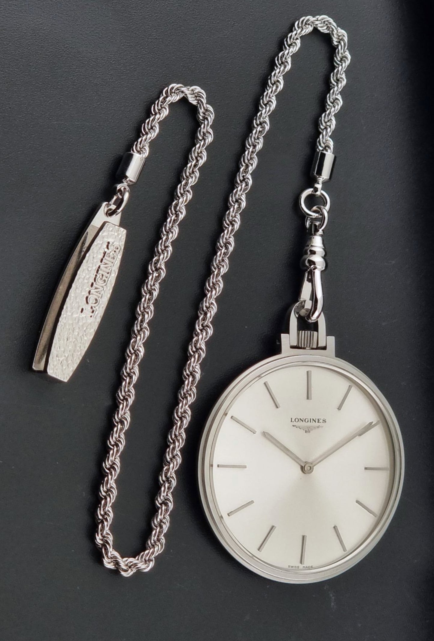 A silver-toned Longines pocket watch with a white dial, displayed alongside its matching braided chain and separate rectangular watch end piece on a dark gray background.