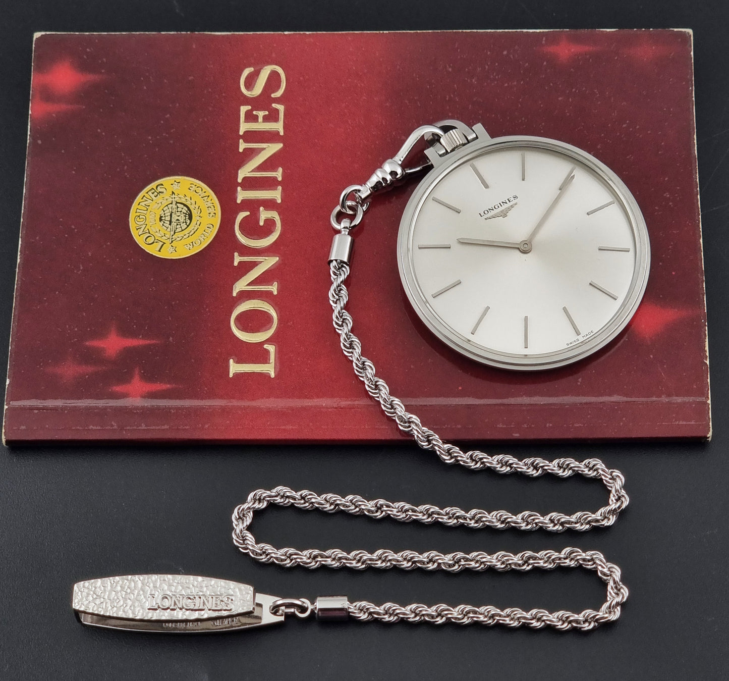 A vintage Longines pocket watch with a silver-toned chain and a white dial, displayed on top of its red booklet with gold Longines lettering.
