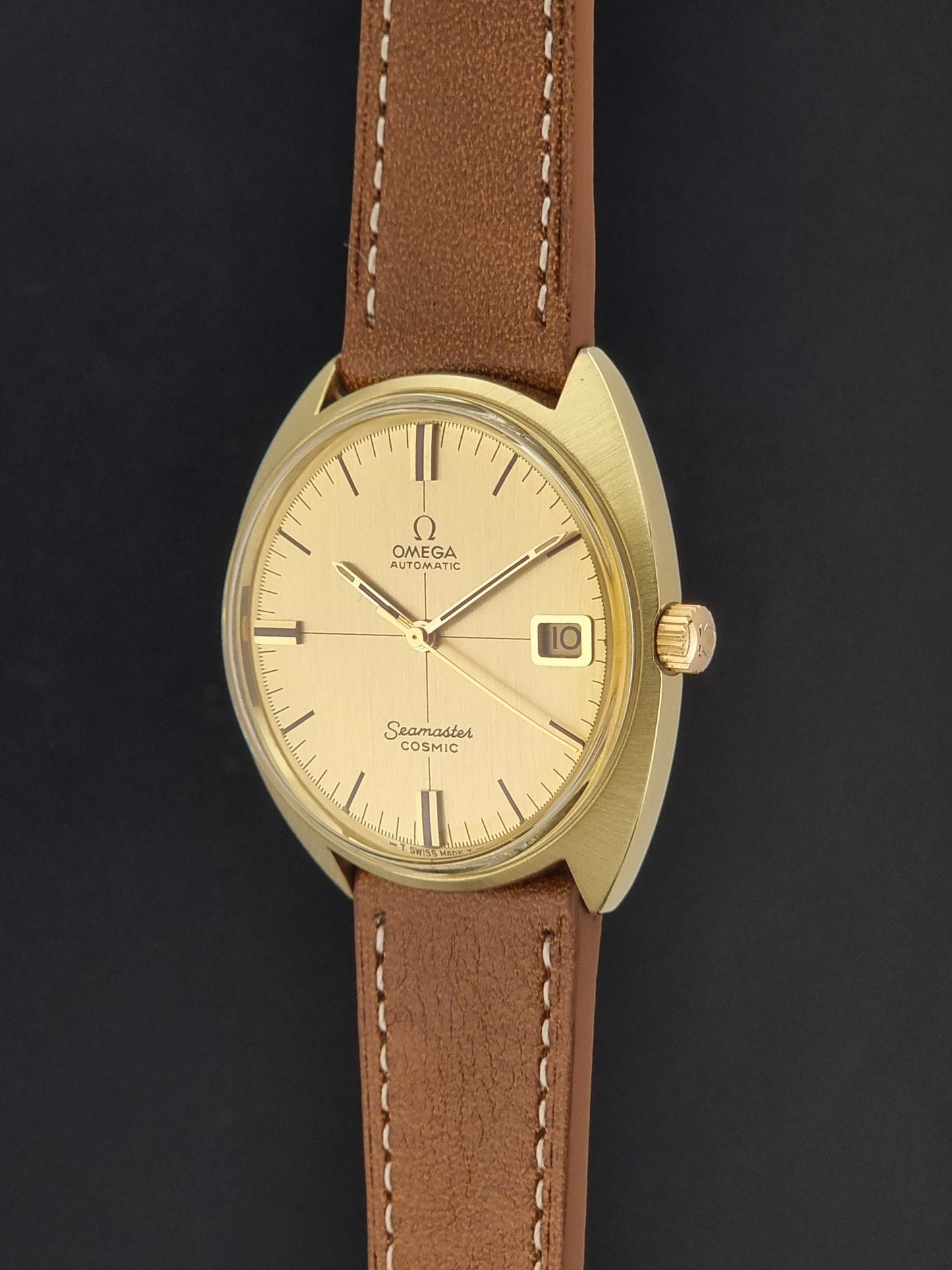An Omega Seamaster Cosmic wristwatch with a gold-tone case, cream-colored dial, and a brown leather strap with white stitching, shown against a dark background.