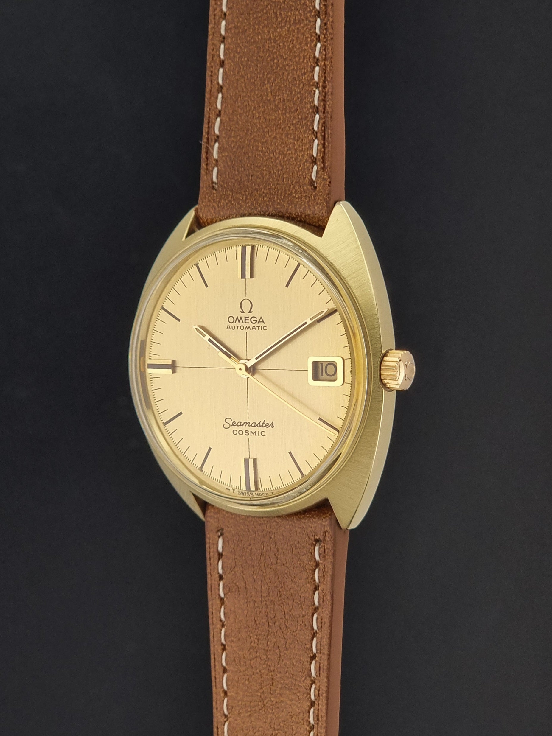 An Omega Seamaster Cosmic wristwatch with a gold-tone case, cream-colored dial, and a brown leather strap with white stitching, shown against a dark background.