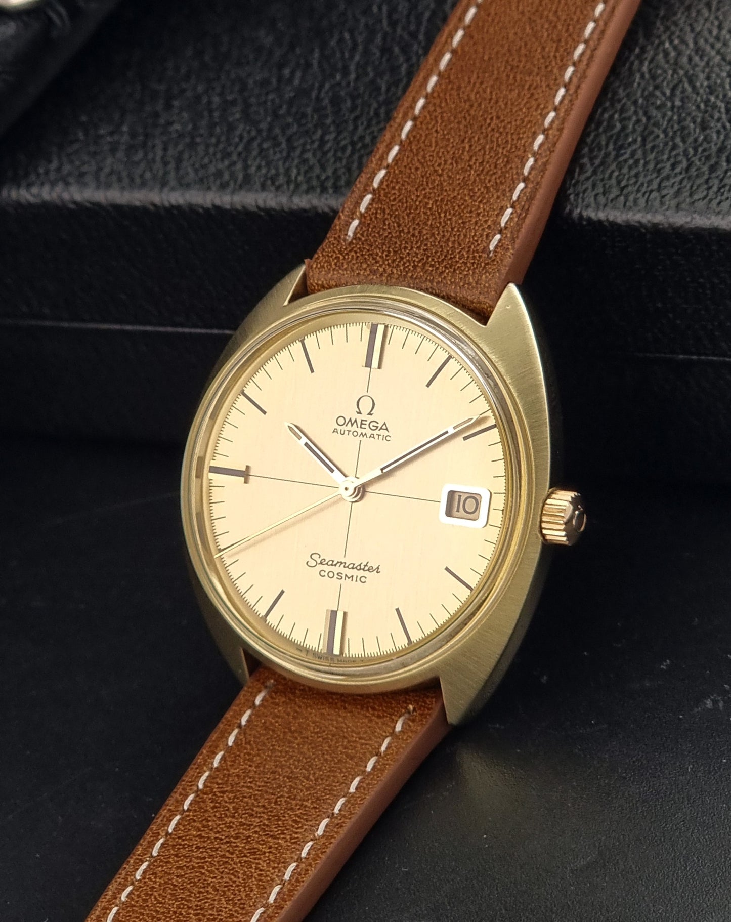 A vintage Omega Seamaster Cosmo wristwatch with a gold-tone case, cream-colored dial, and a brown leather strap with white stitching, resting on a black textured surface.