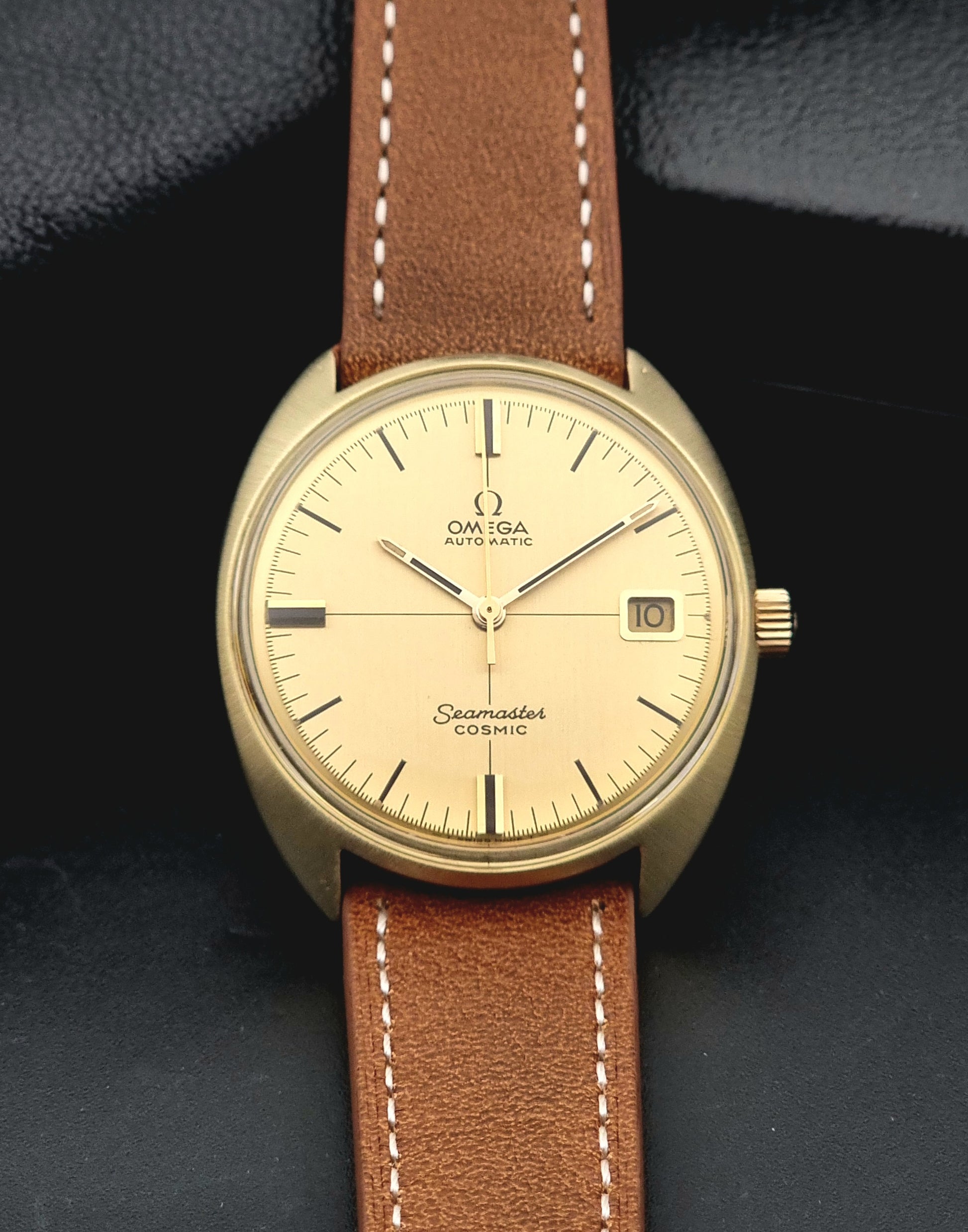 An Omega Seamaster Cosmograph wristwatch with a round gold-tone case, cream-colored dial, and a brown leather strap with white stitching, shown against a dark textured background.