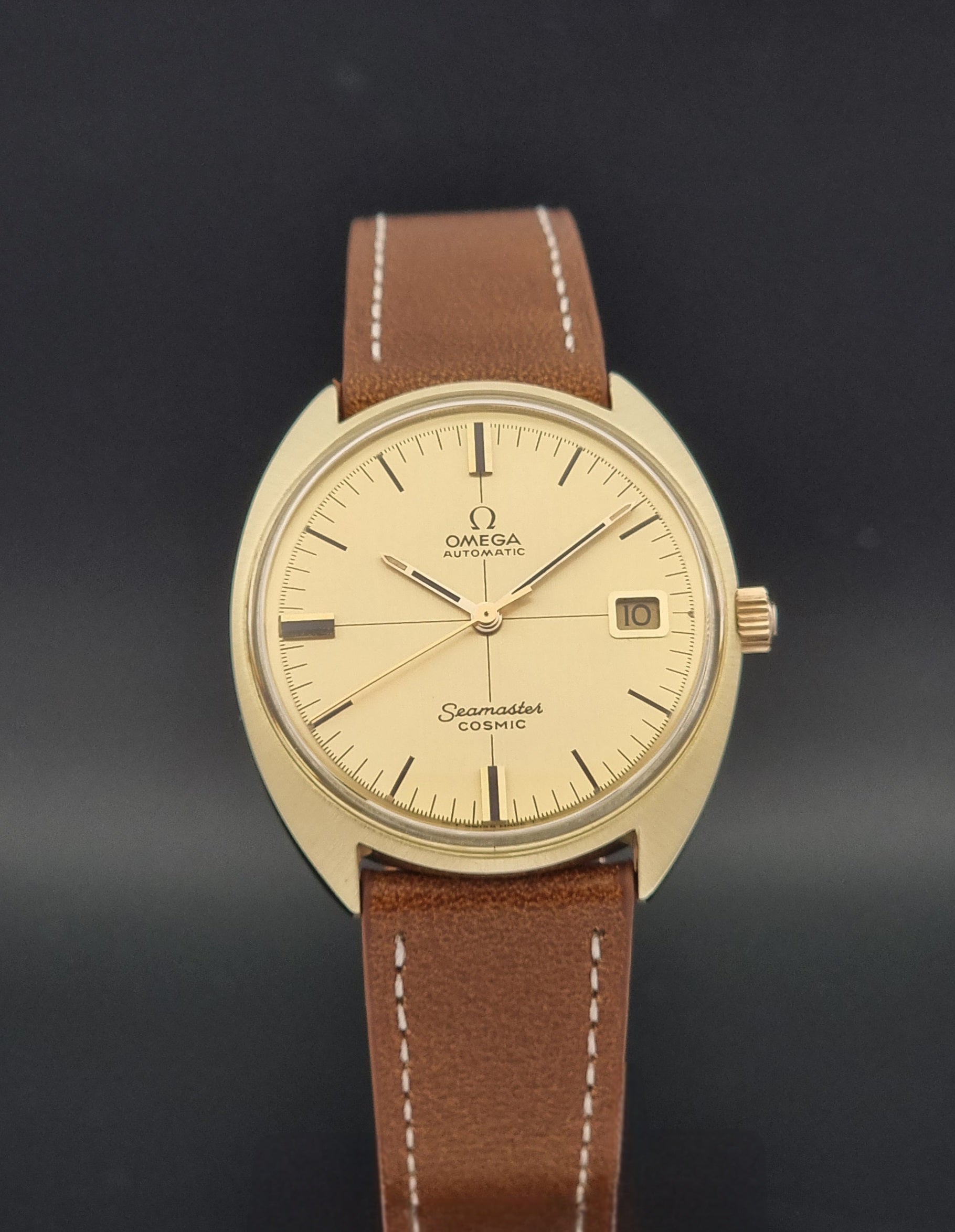 Omega Seamaster Cosmic wristwatch with a gold-tone case, cream dial, and brown leather strap with white stitching, shown against a dark gray background.