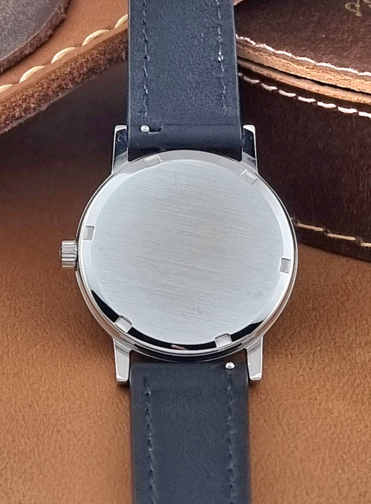 Close-up of the back of a A Vintage Omega Geneve 136.0098 watch, and black leather strap, resting on a brown leather surface.