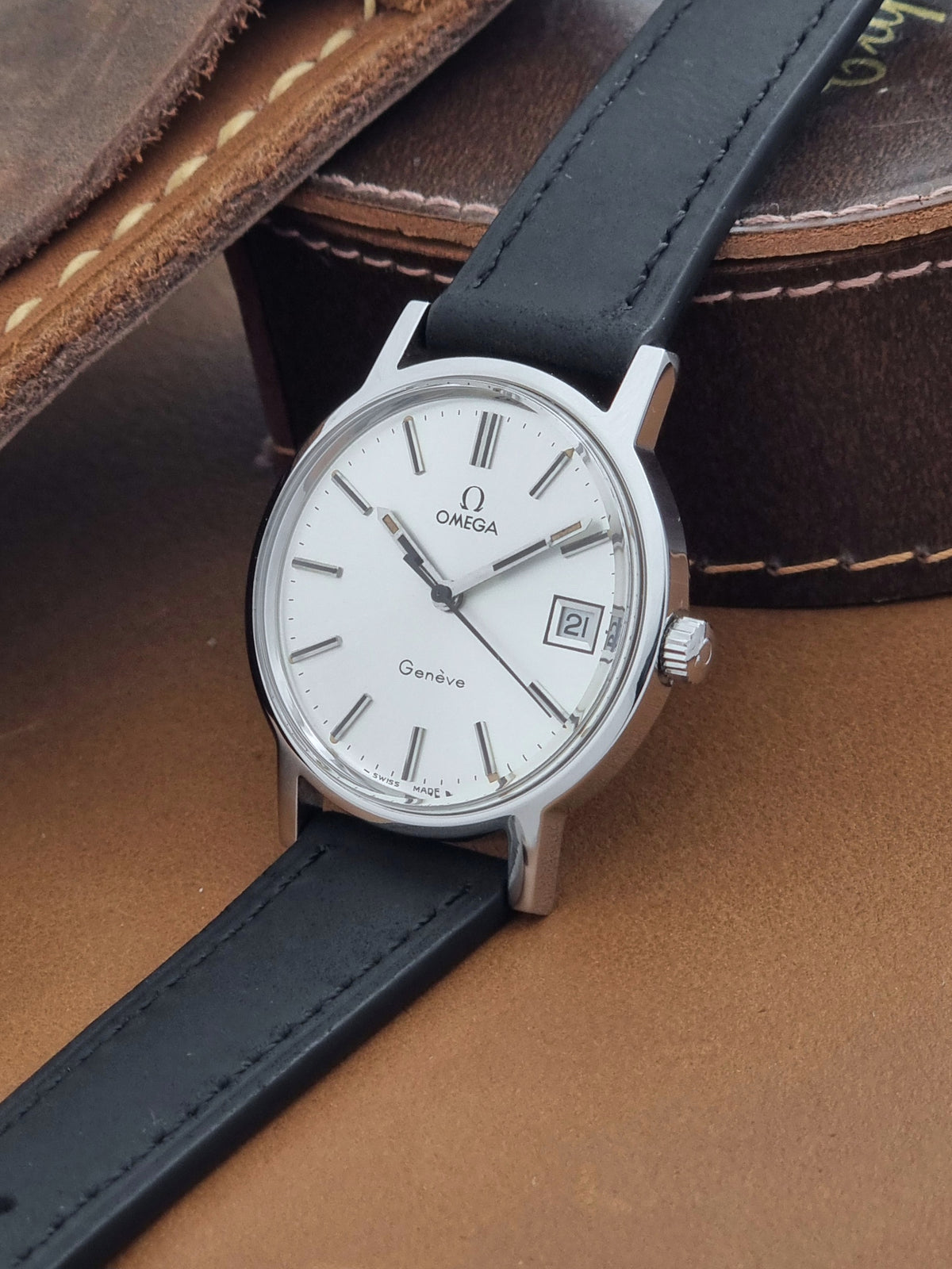 A Vintage Omega Geneve 136.0098 watch, white dial, and black leather strap, resting on a brown leather surface.