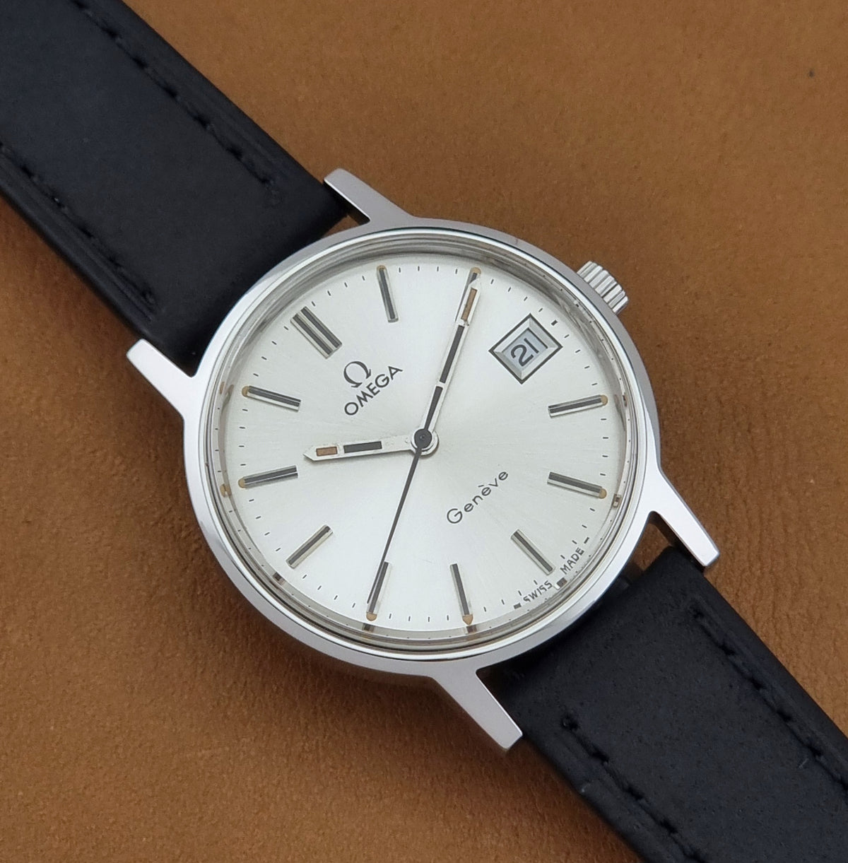 A Vintage Omega Geneve 136.0098 watch, white dial, and black leather strap shown diagonally on a textured brown background.