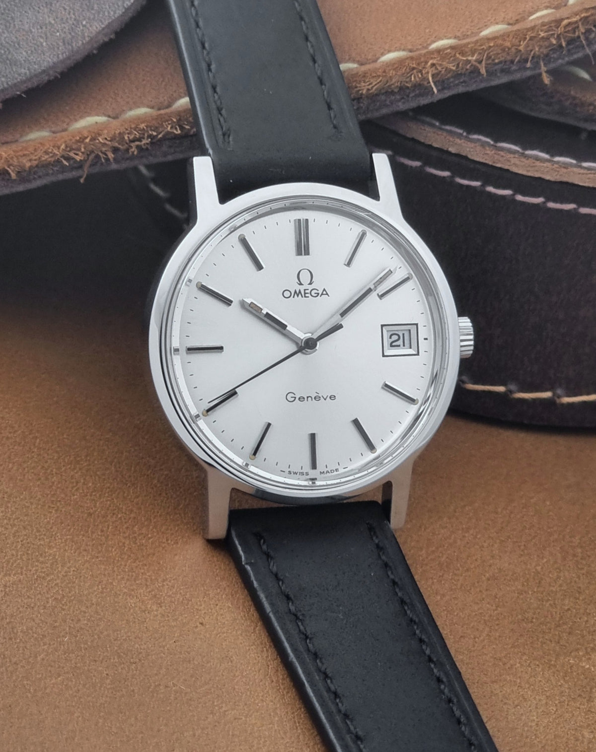 A Vintage Omega Geneve 136.0098 watch, white dial, and black leather strap, resting on a brown leather surface.