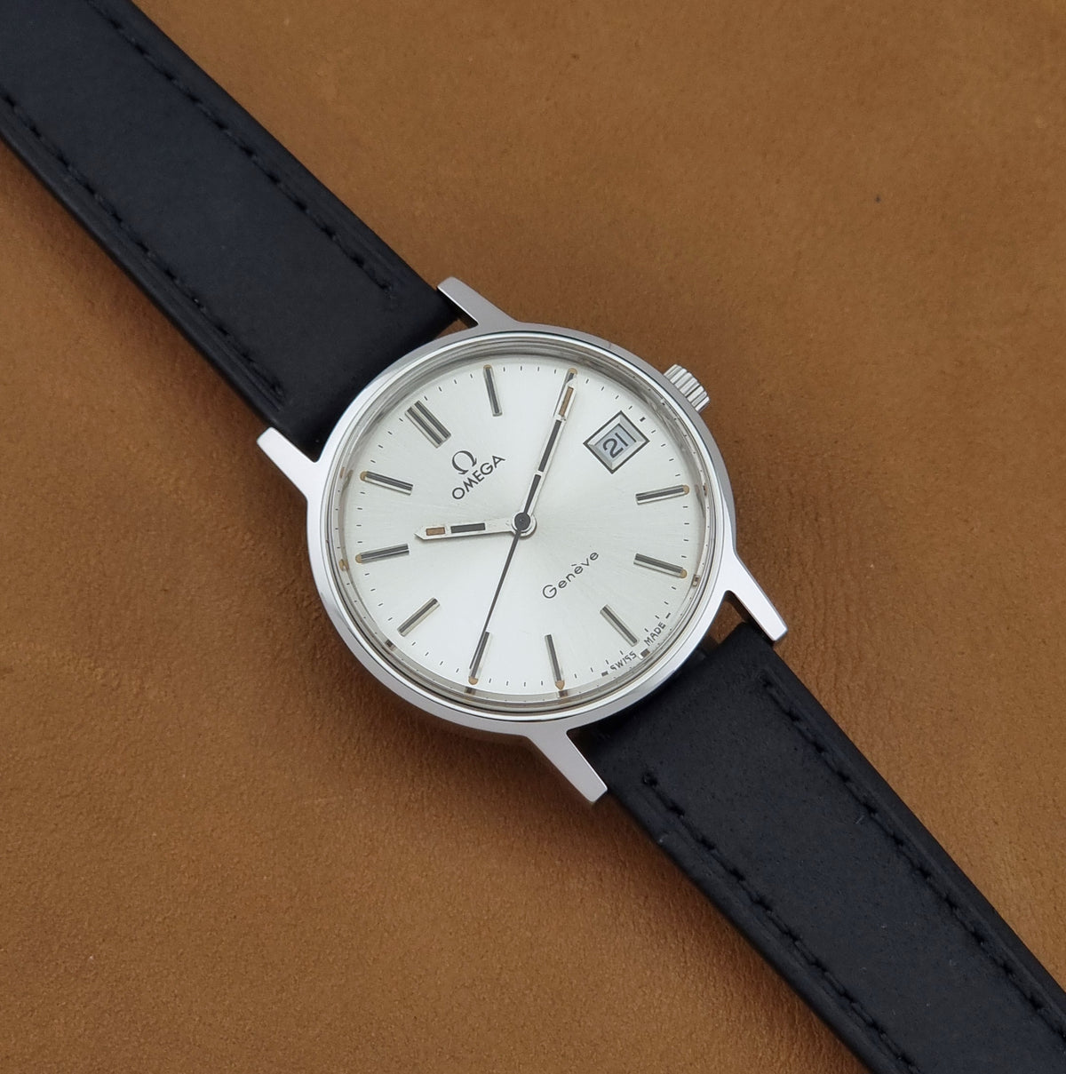 A Vintage Omega Geneve 136.0098 watch, white dial, and black leather strap, shown diagonally on a brown textured background.