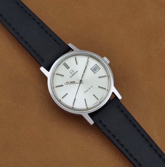 A Vintage Omega Geneve 136.0098 watch, white dial, and black leather strap, shown diagonally on a brown textured background.