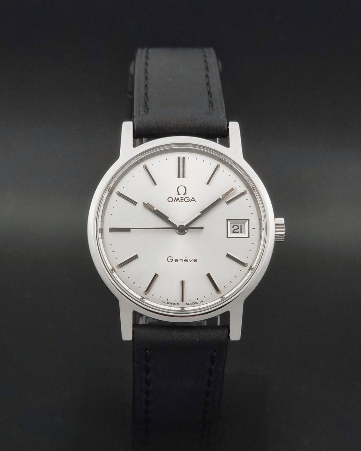A Vintage Omega Geneve 136.0098 watch, white dial, and black leather strap, photographed against a dark gray background.